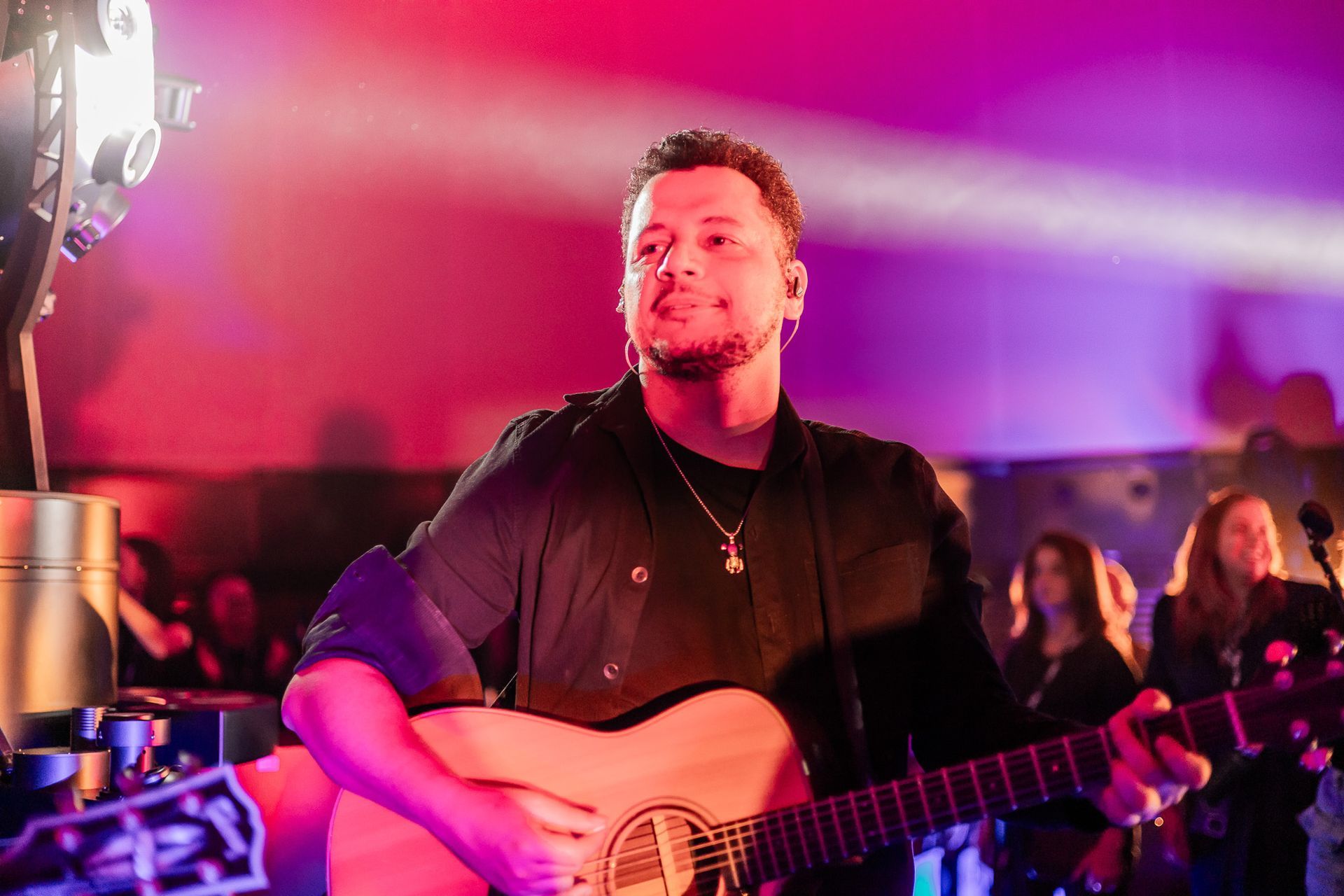 Um músico toca violão no palco sob iluminação roxa, com uma banda e o público visíveis ao fundo.