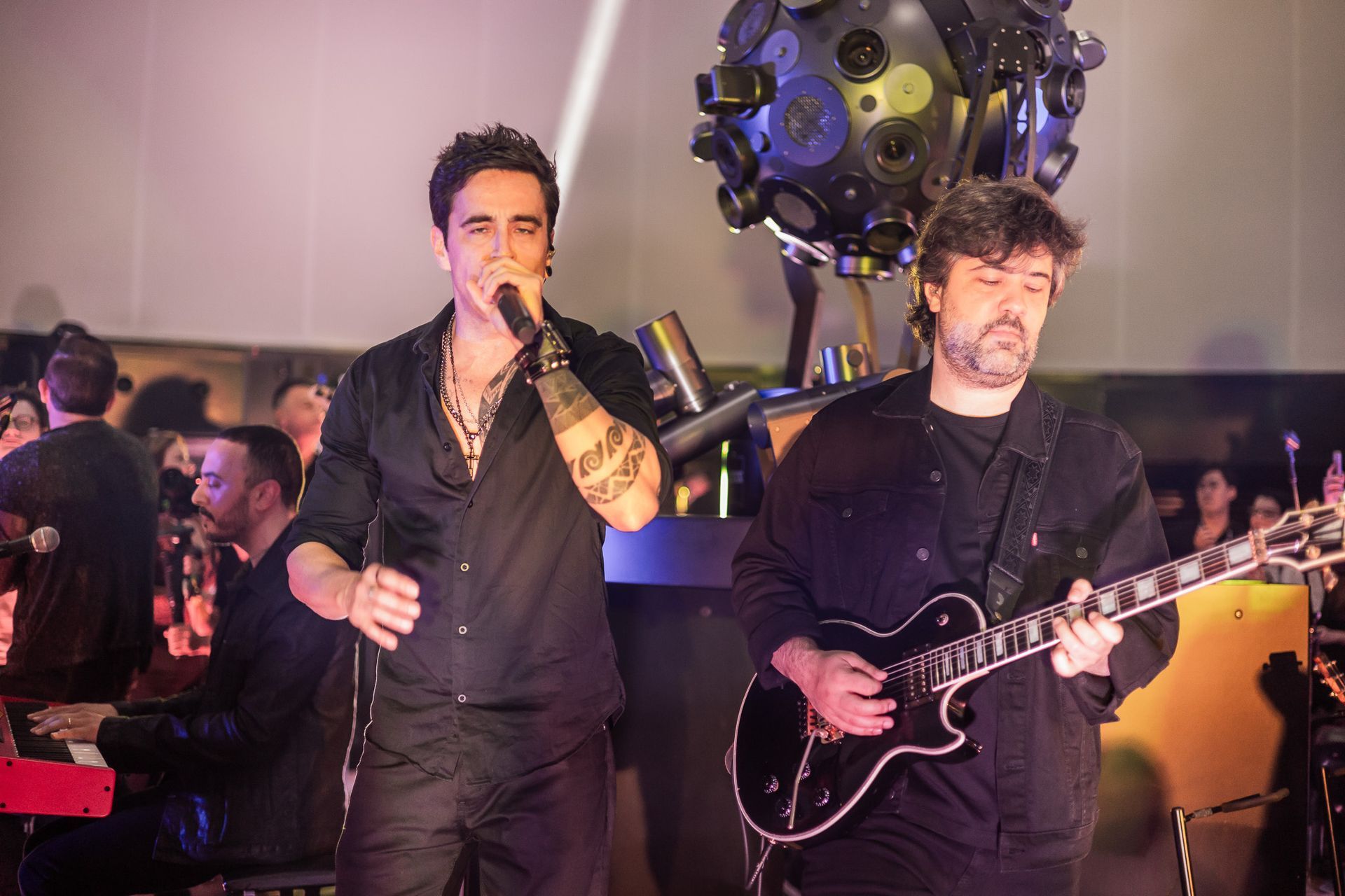 Um vocalista se apresenta com um microfone enquanto um guitarrista toca próximo a ele, ambos vestidos de escuro em um palco.