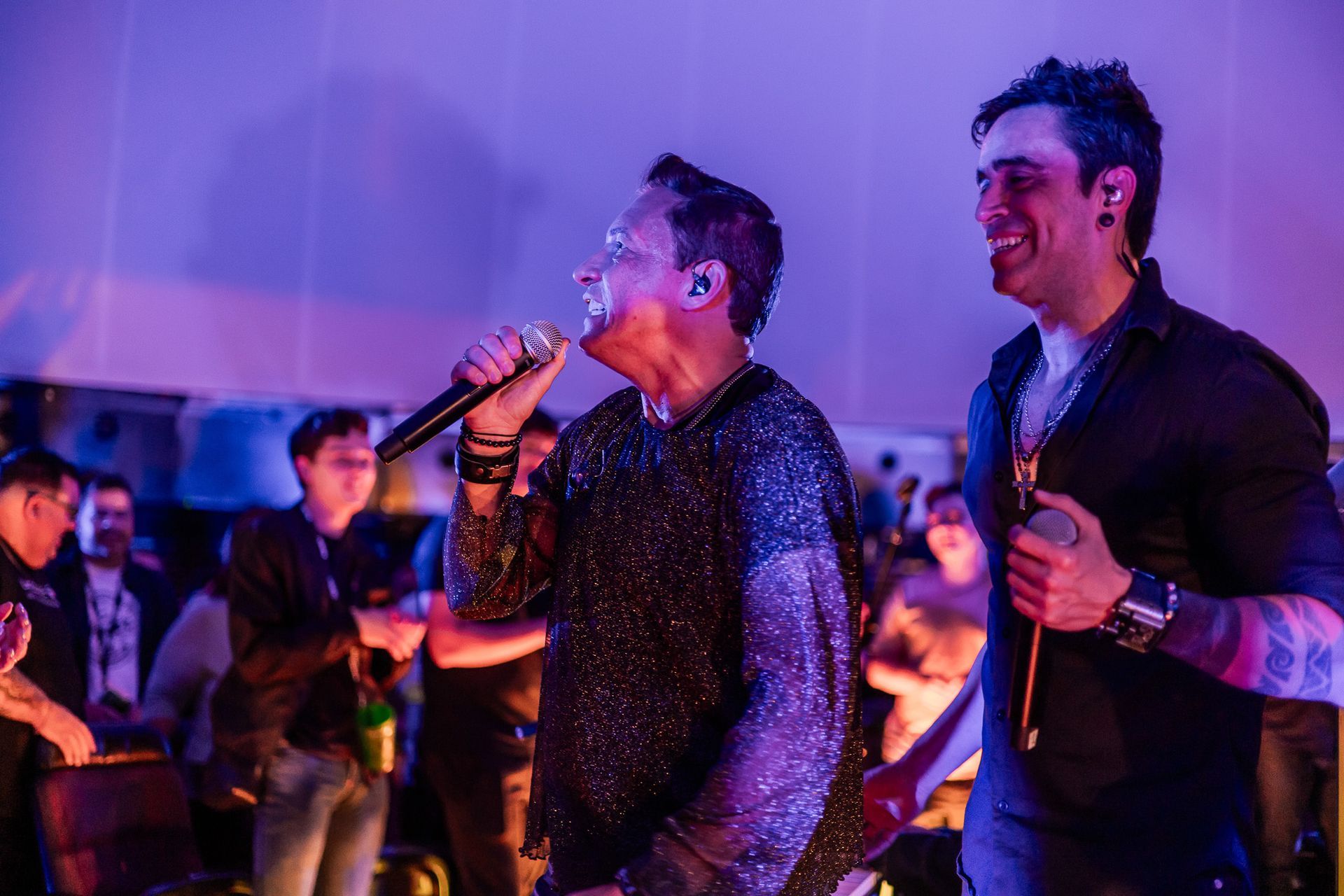 Dois músicos se apresentam no palco sob luzes roxas, um cantando ao microfone e o outro sorrindo ao seu lado.