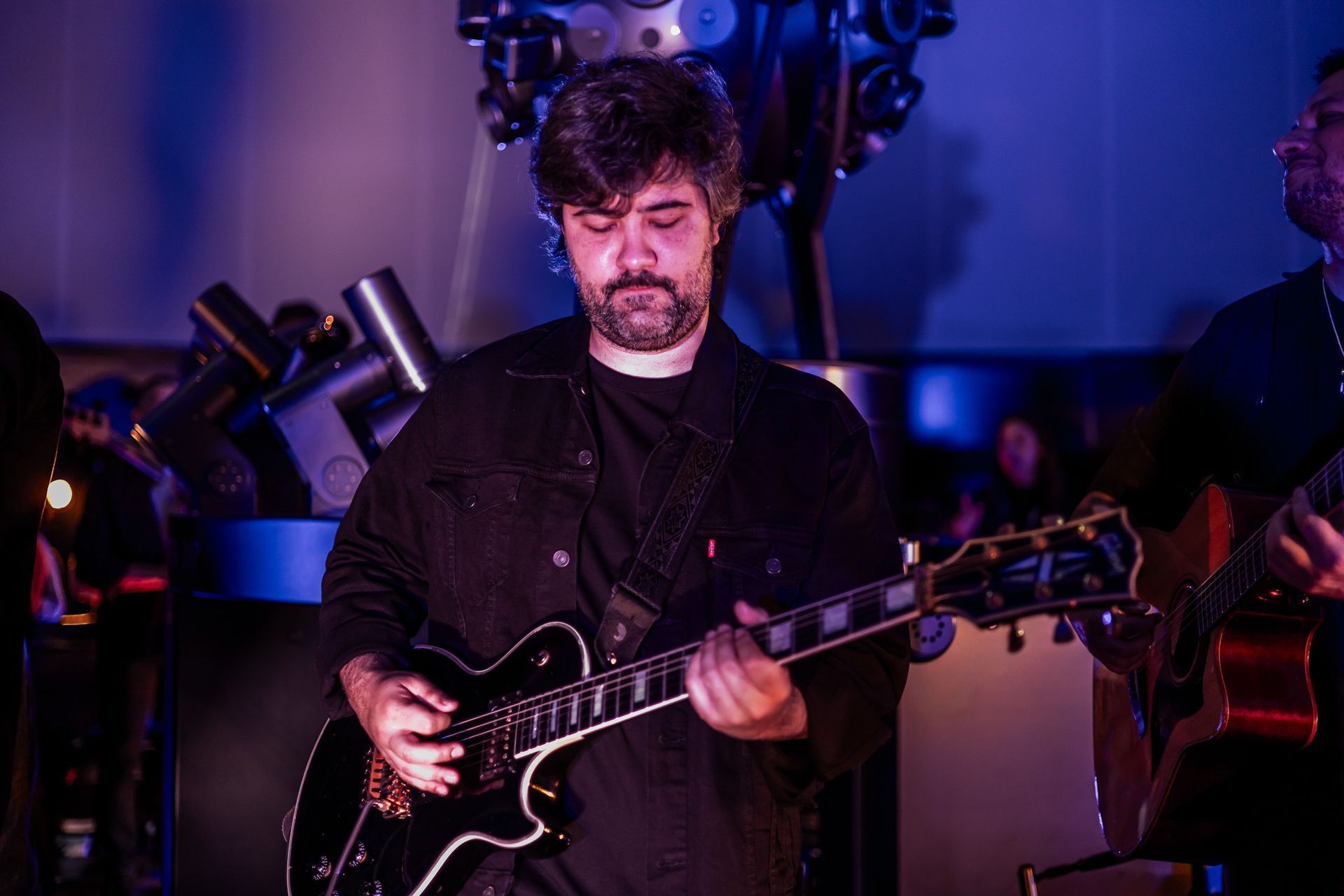 Um artista com barba toca uma guitarra elétrica preta em um palco iluminado com tons de azul.