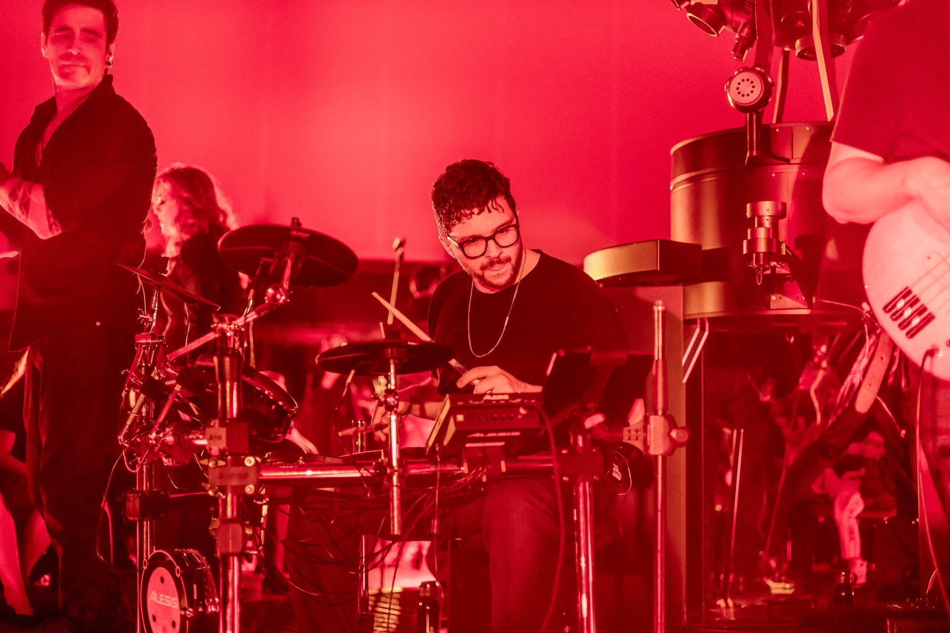 Uma banda se apresenta no palco sob luzes vermelhas brilhantes, com um baterista e um guitarrista, em um show ao vivo.