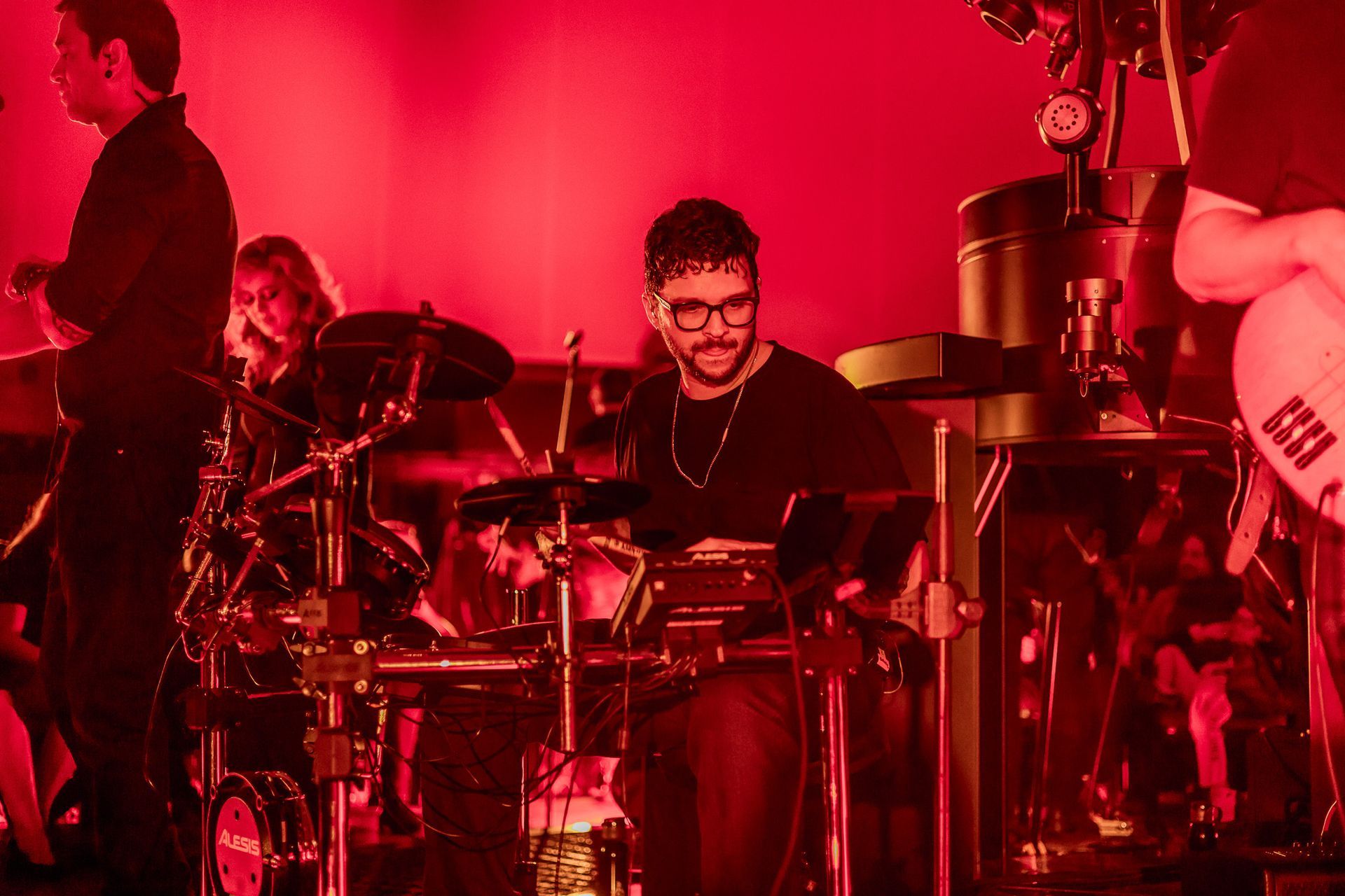 Uma banda se apresenta em um ambiente escuro banhado por luz vermelha, com um baterista concentrado em tocar.
