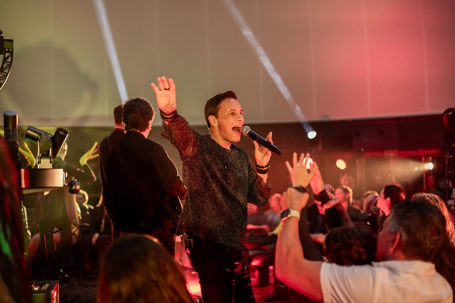 Um artista com uma blusa brilhante canta ao microfone, acenando para a plateia durante um show.