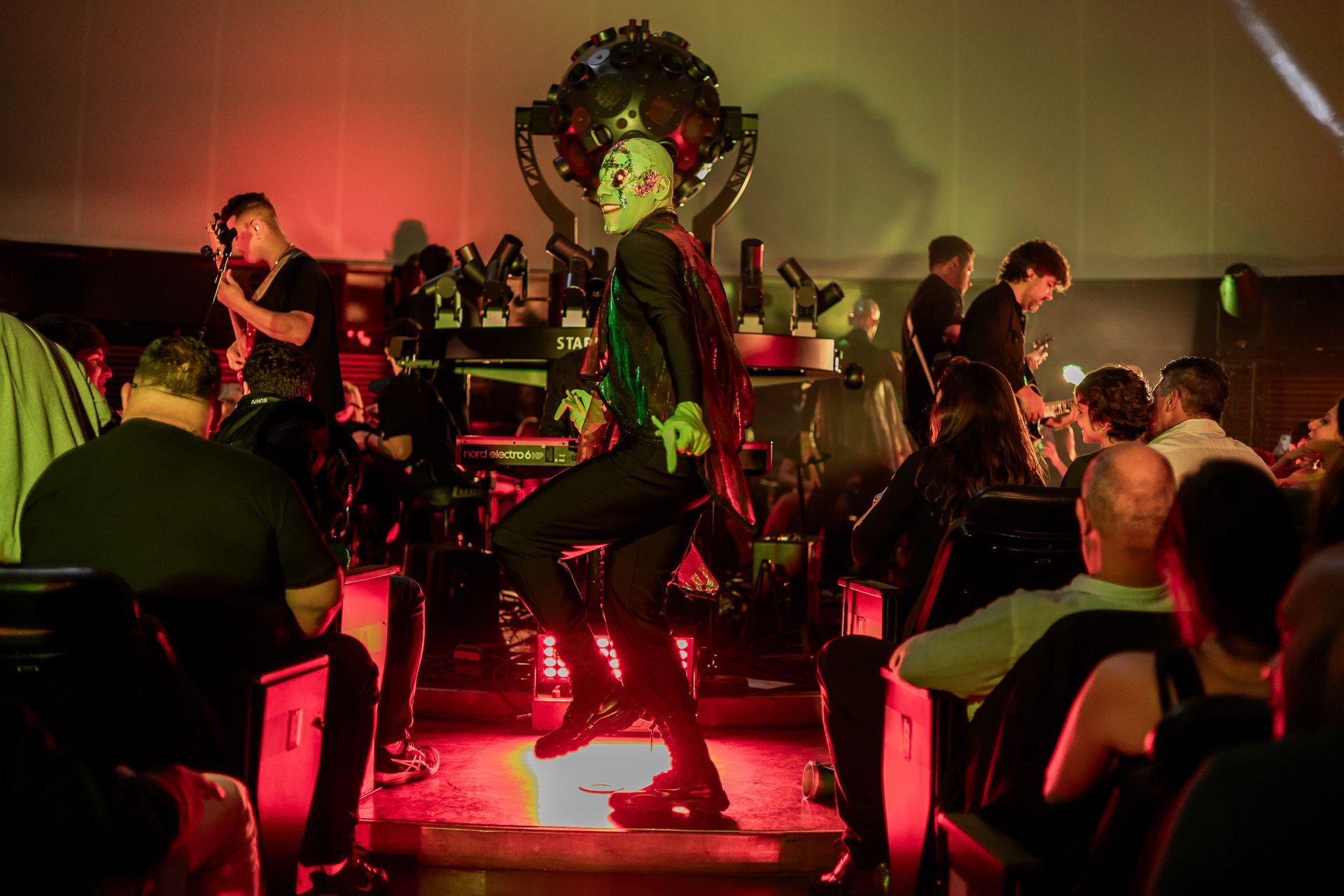 Um artista mascarado e com uma capa escura dança em um palco bem iluminado diante de uma plateia com pouca luz.