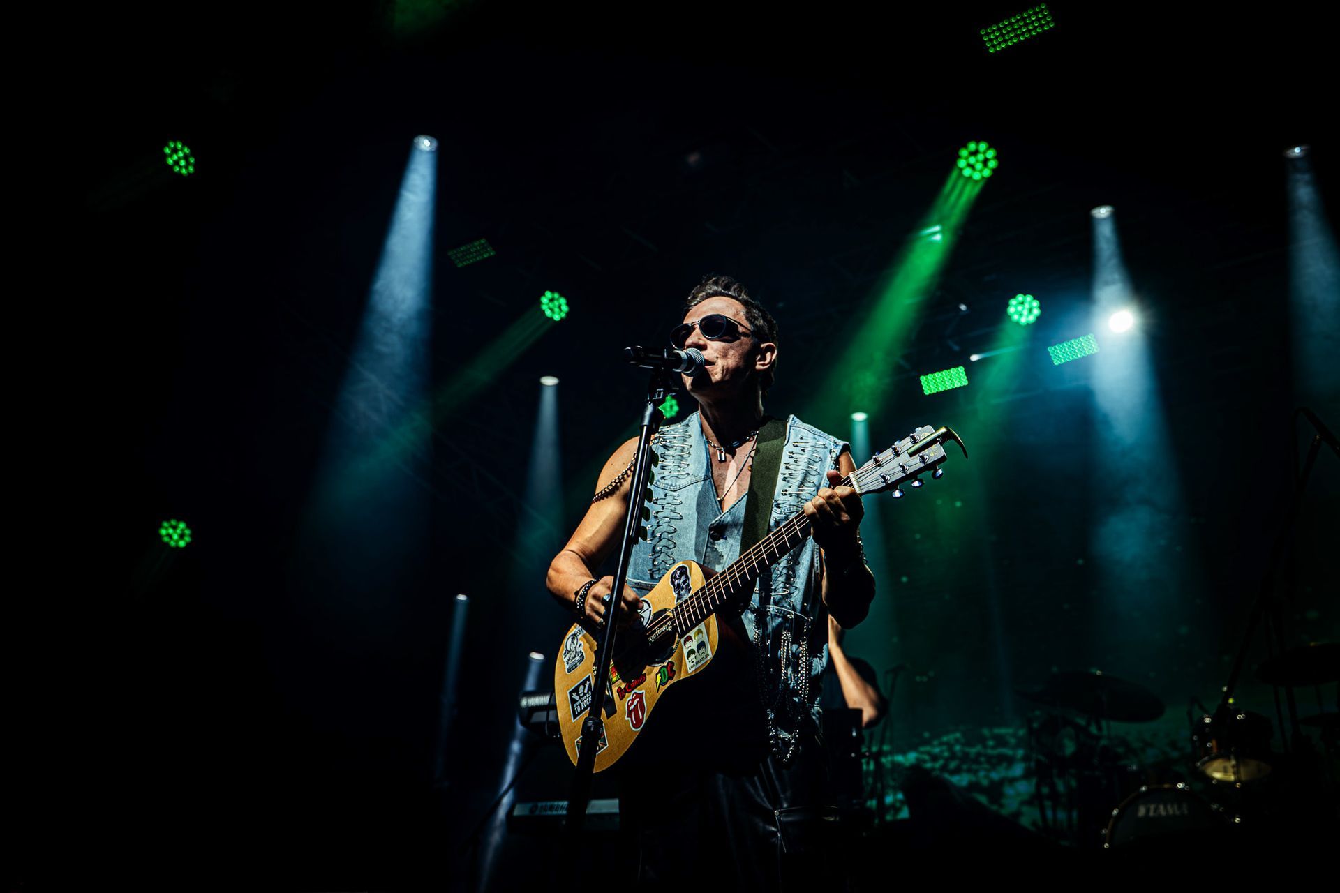 Um músico de óculos escuros toca guitarra no palco, iluminado por holofotes verdes e azuis.
