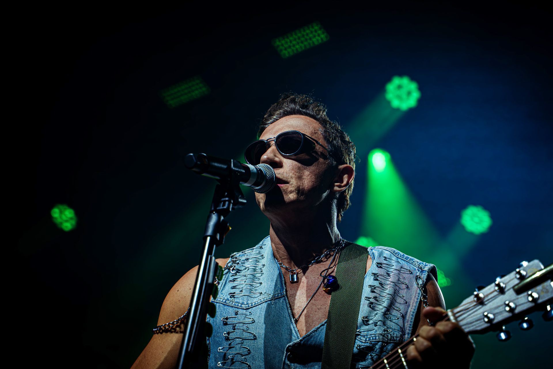 Um musicista usando óculos escuros e uma blusa sem mangas bordada se apresenta no palco sob holofotes verdes.