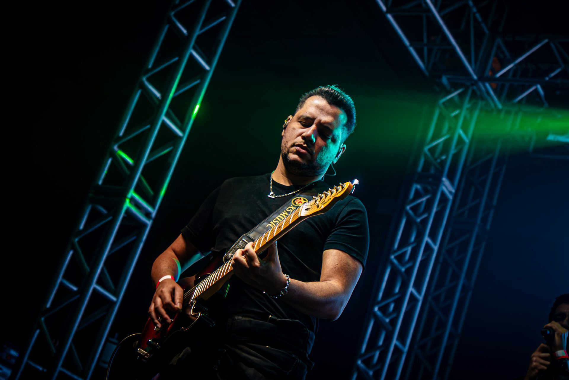 Um músico de camiseta preta toca uma guitarra elétrica com um padrão intrincado no braço sob luzes de palco verde-brilhantes.