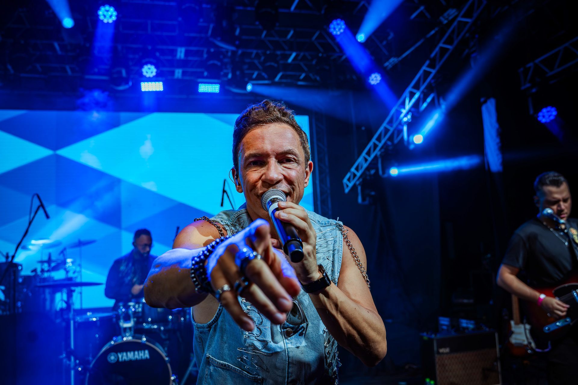 Um artista cantando em um palco com iluminação azul, estendendo a mão em direção à câmera com uma banda ao fundo.