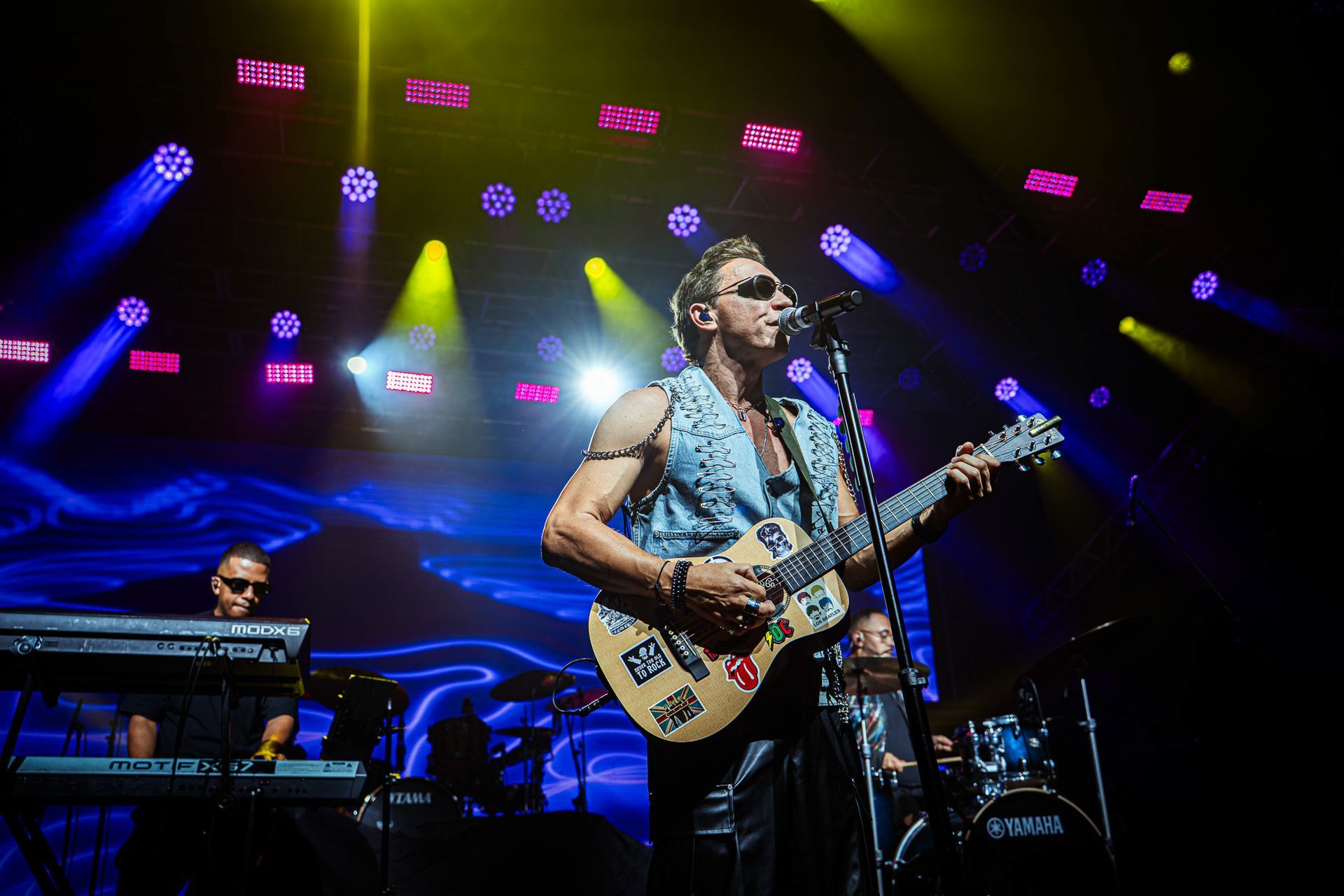 Um músico de óculos escuros e colete sem mangas toca violão em um palco de concertos iluminado por luzes azuis e amarelas.