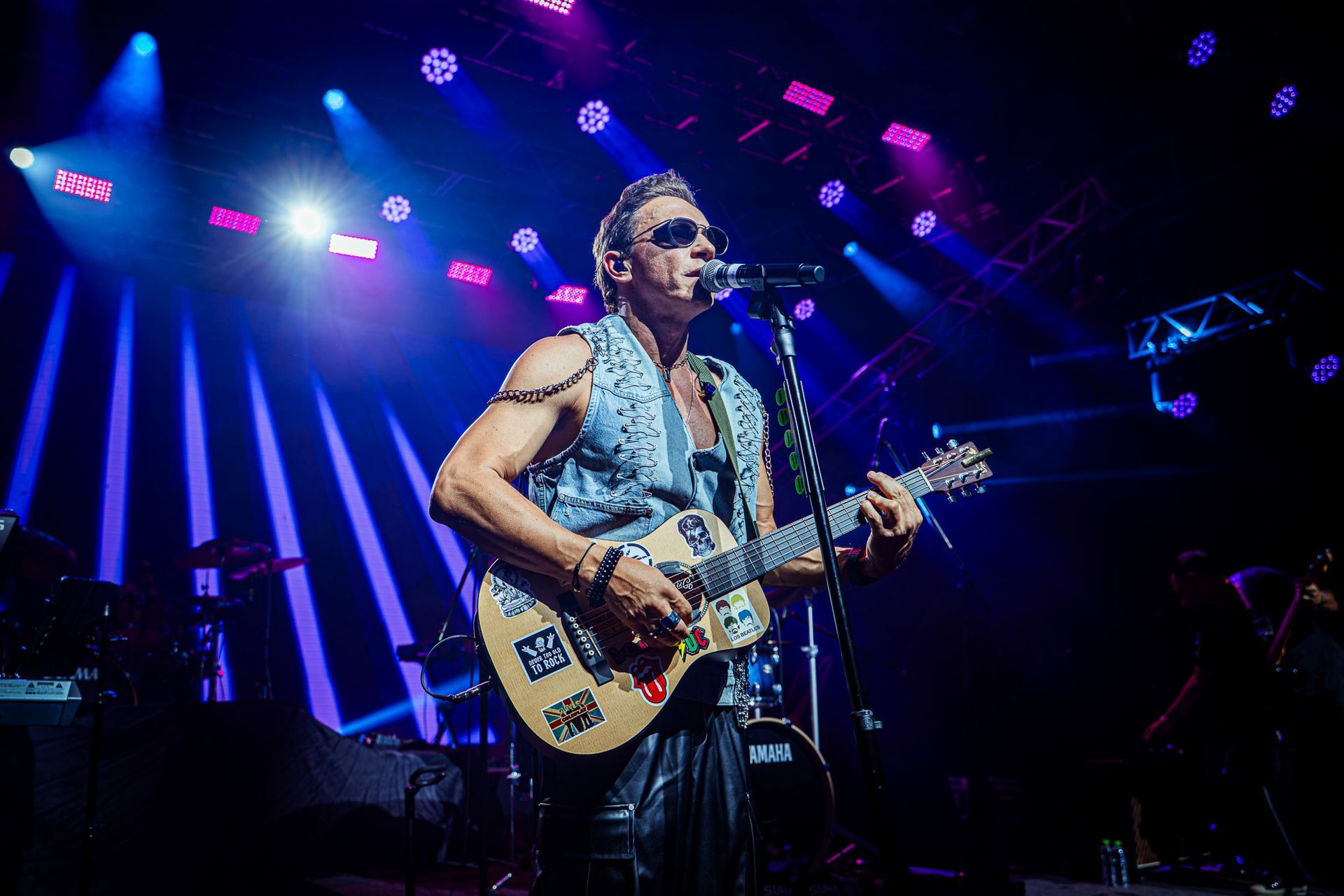 Um músico com um colete cravejado de tachas toca um violão decorado enquanto canta ao microfone em um palco de concerto.