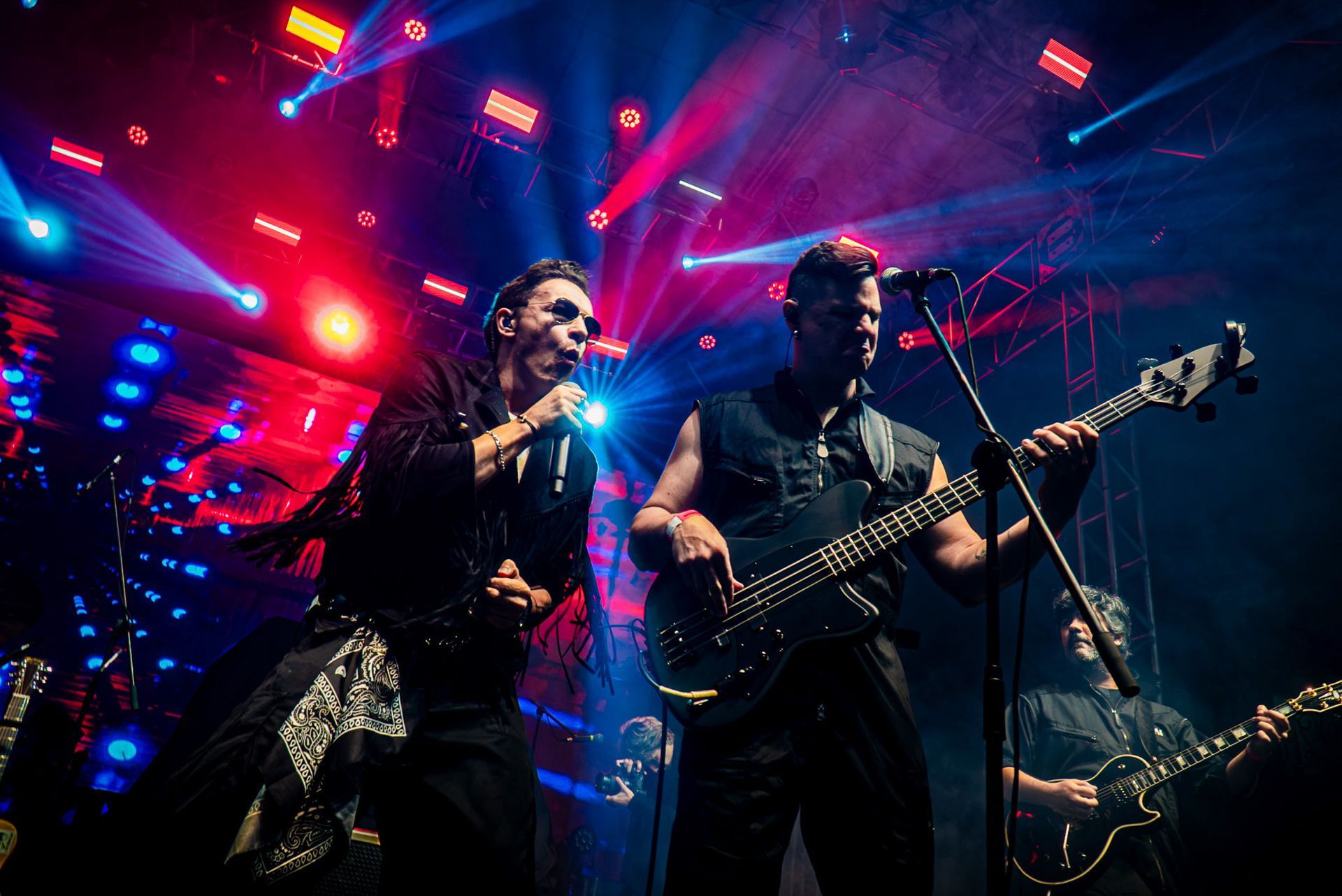 Dois artistas em um palco com iluminação vibrante em vermelho e azul; um canta ao microfone, o outro toca baixo.