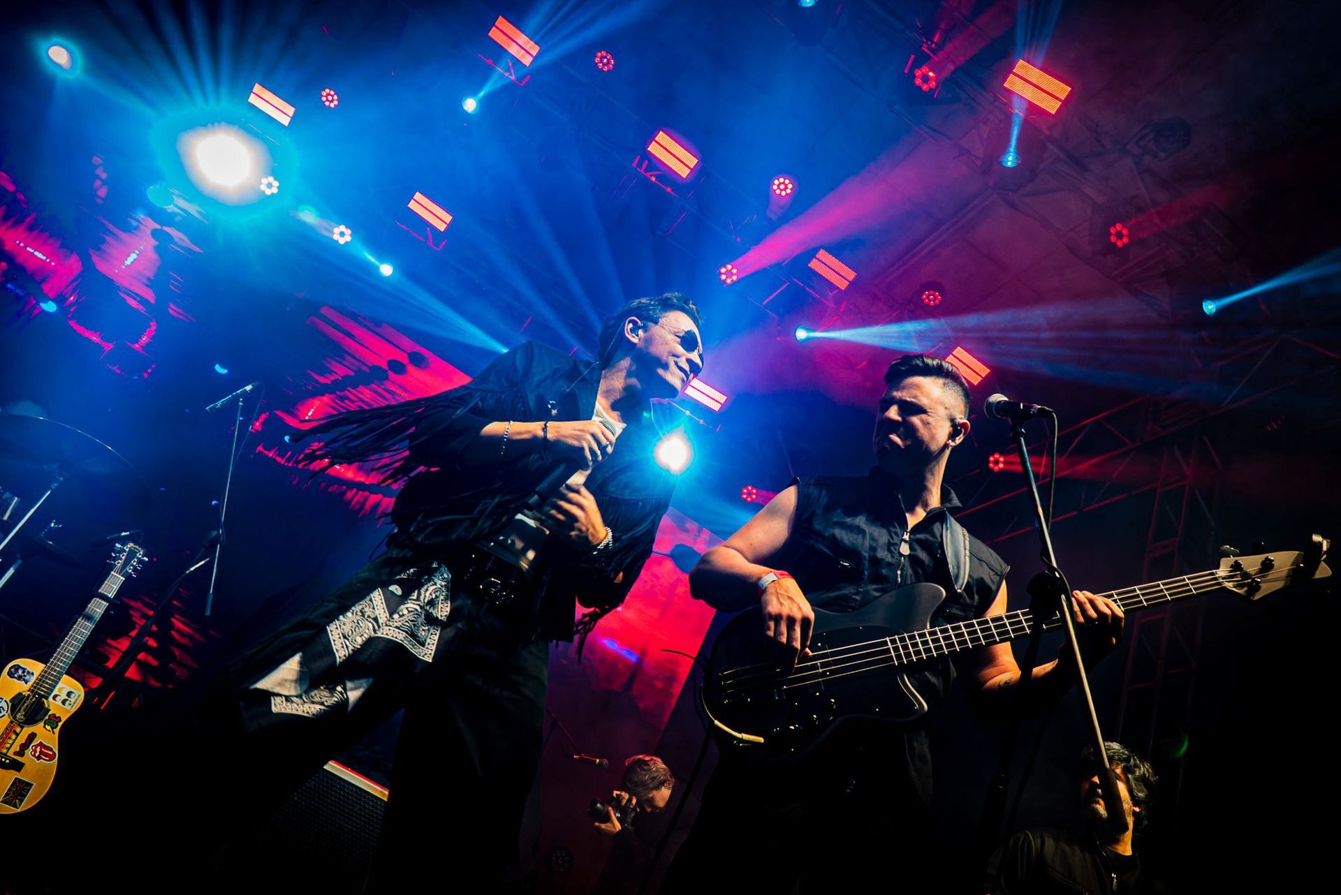 Dois músicos se apresentam em um palco, um cantando ao microfone e o outro tocando baixo.