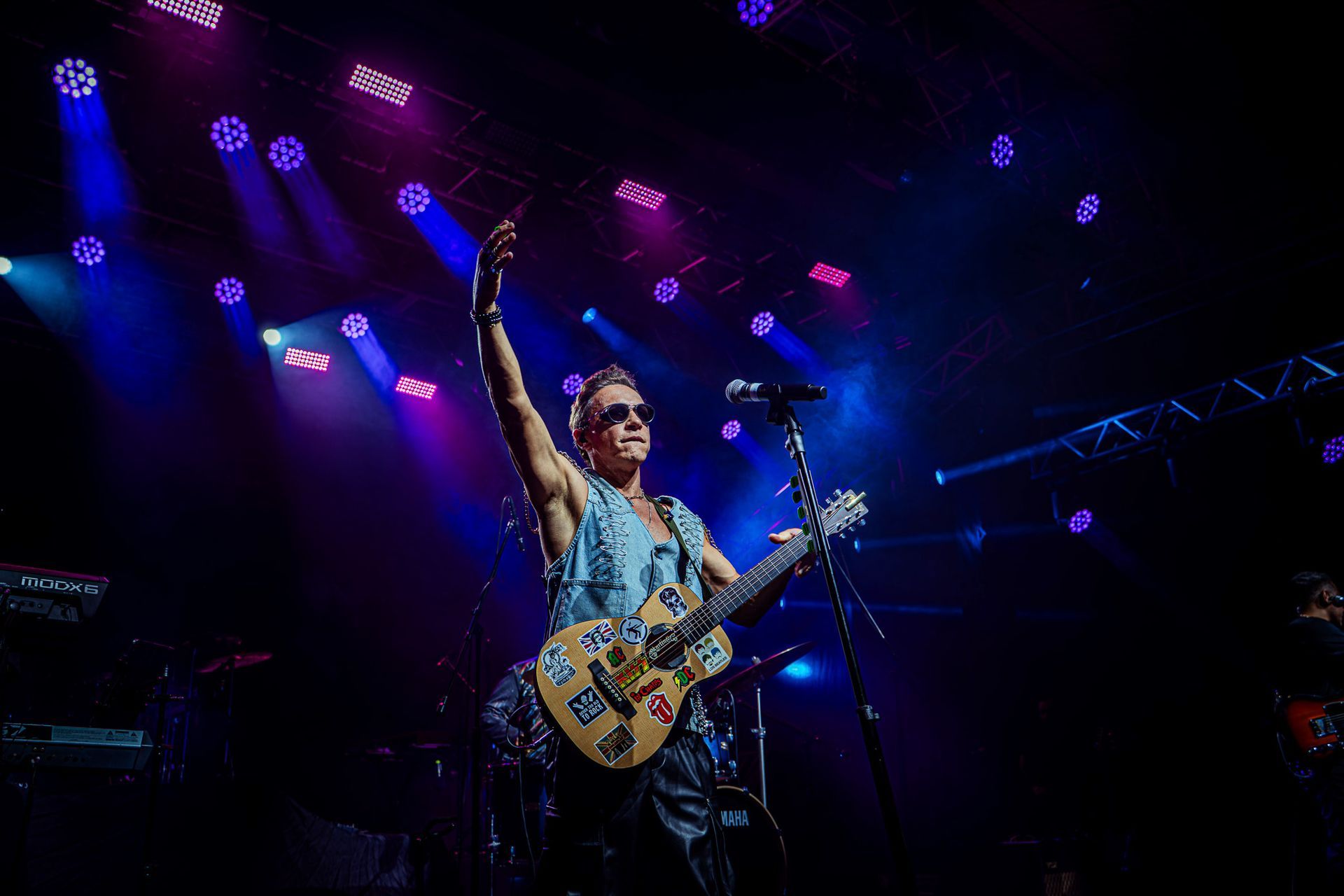 Um músico de óculos escuros tocando uma guitarra elétrica coberta de adesivos e levantando o braço no palco sob luzes azuis.