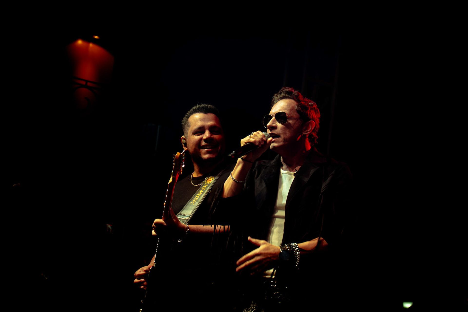 Dois músicos se apresentando em um palco escuro, um cantando ao microfone e o outro segurando uma guitarra elétrica.