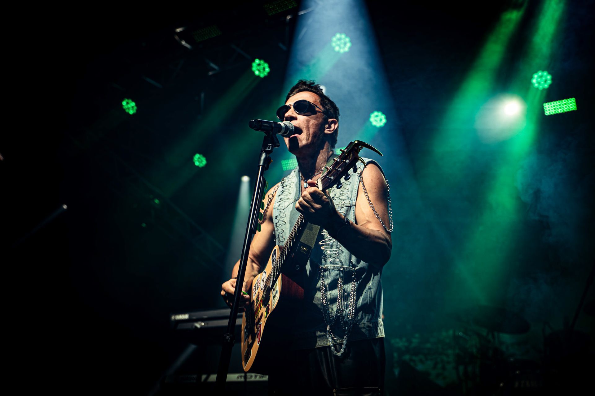 Um músico de óculos escuros, tocando guitarra e cantando no palco sob luzes verdes.