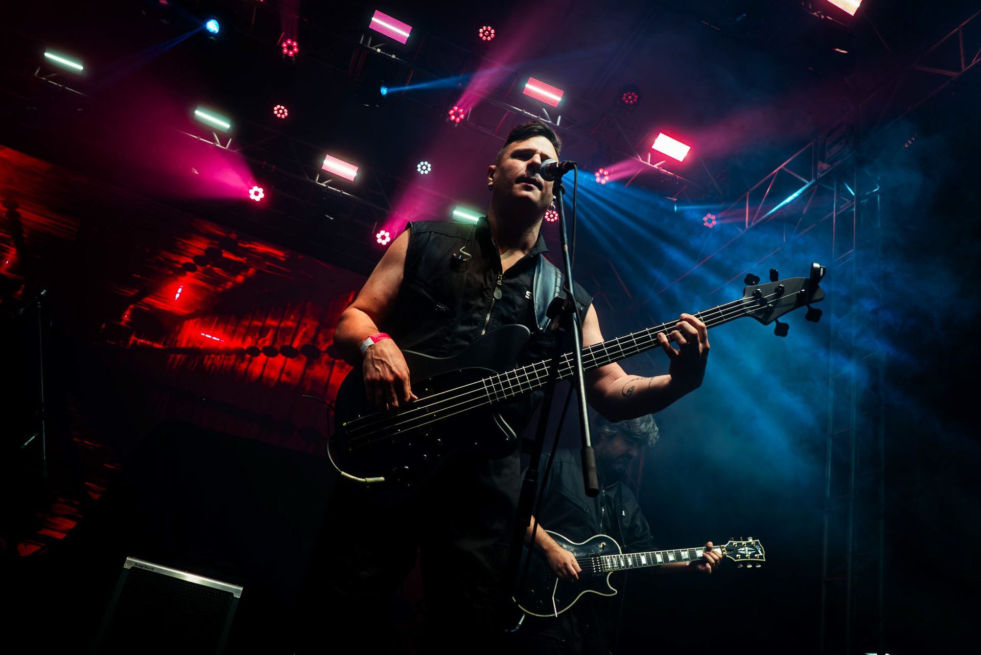 Um artista toca baixo enquanto canta ao microfone em um palco iluminado por holofotes vermelhos e azuis.