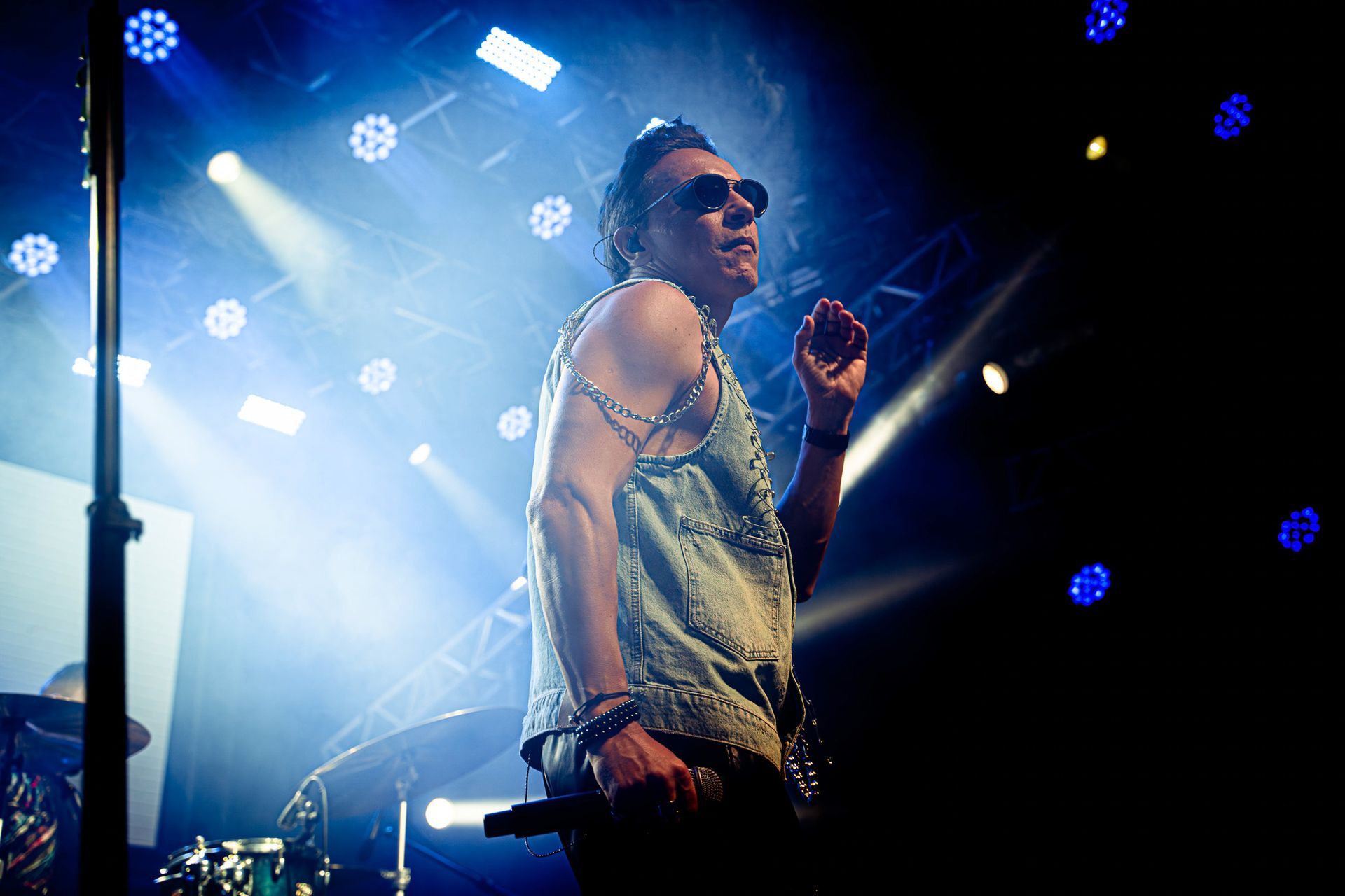 Um artista de óculos escuros e colete sem mangas está em um palco iluminado por holofotes brilhantes, segurando um microfone.