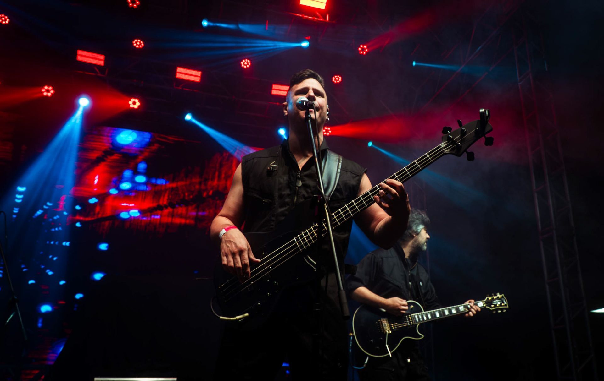 Um músico canta enquanto toca baixo em um palco de concerto com iluminação fraca, em tons de azul e vermelho.