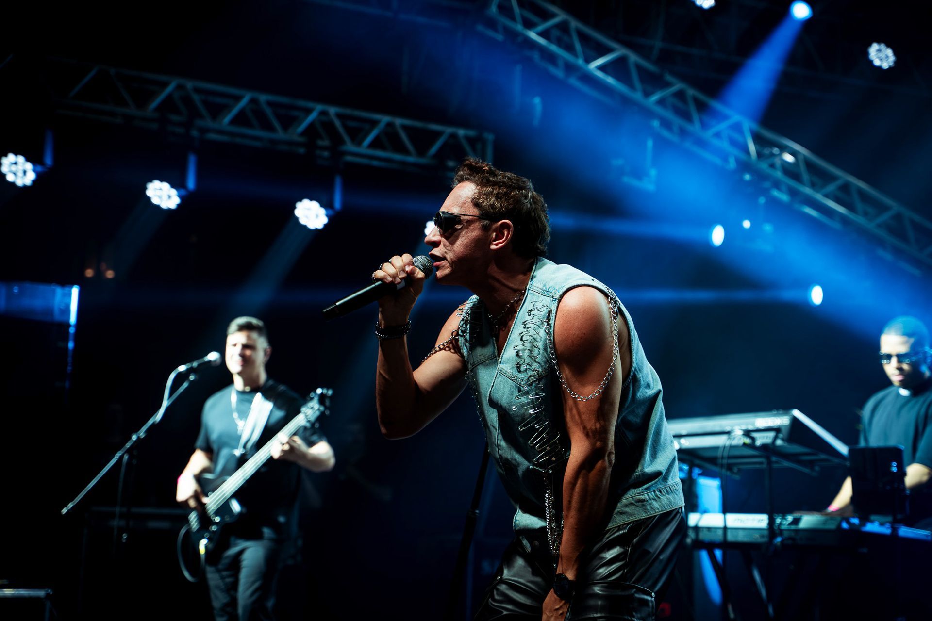 O vocalista principal se apresenta no palco, acompanhada por um músico e um tecladista sob luzes artificiais.