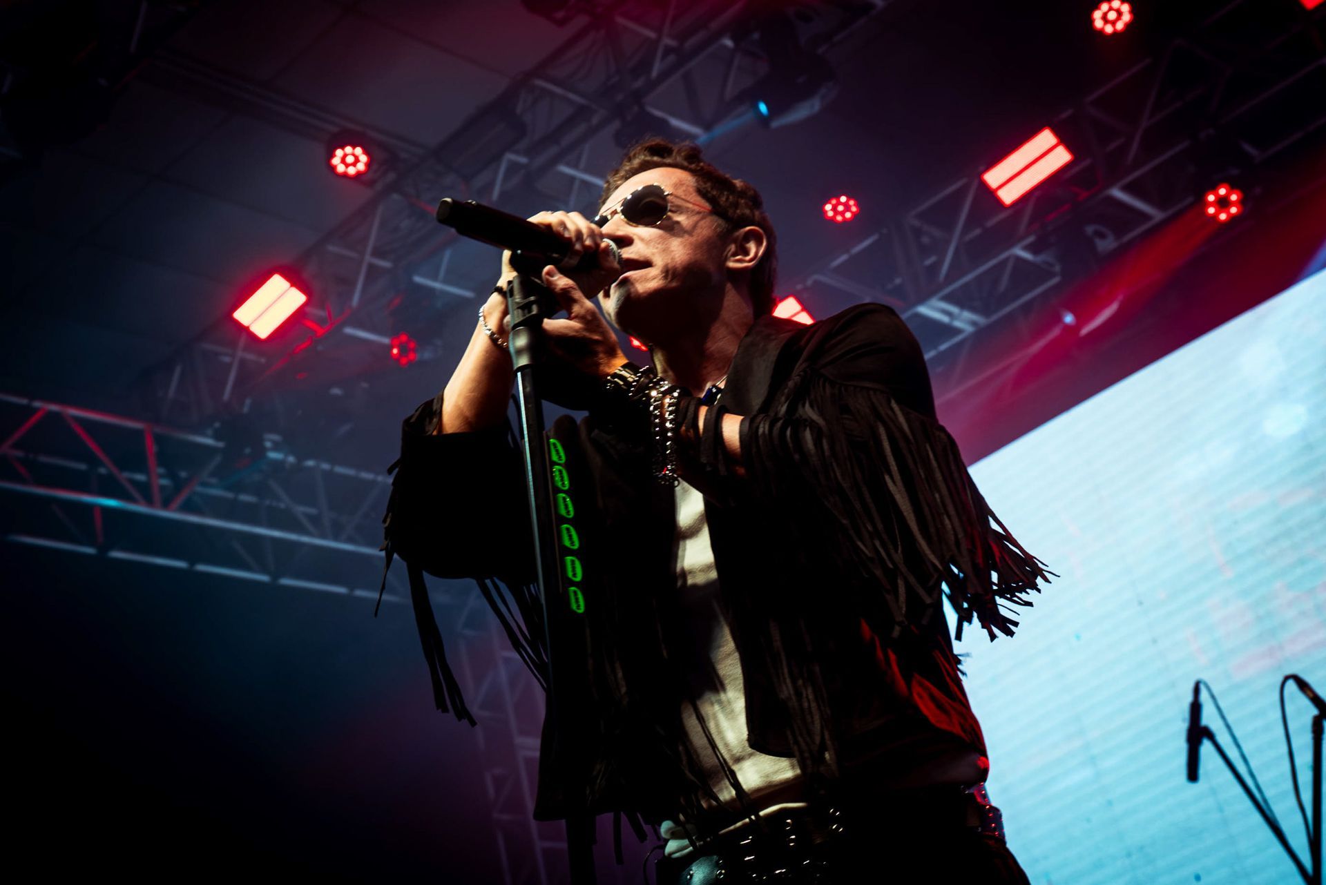 Um artista vestindo uma jaqueta preta com franjas canta em um microfone em um palco com holofotes vermelhos