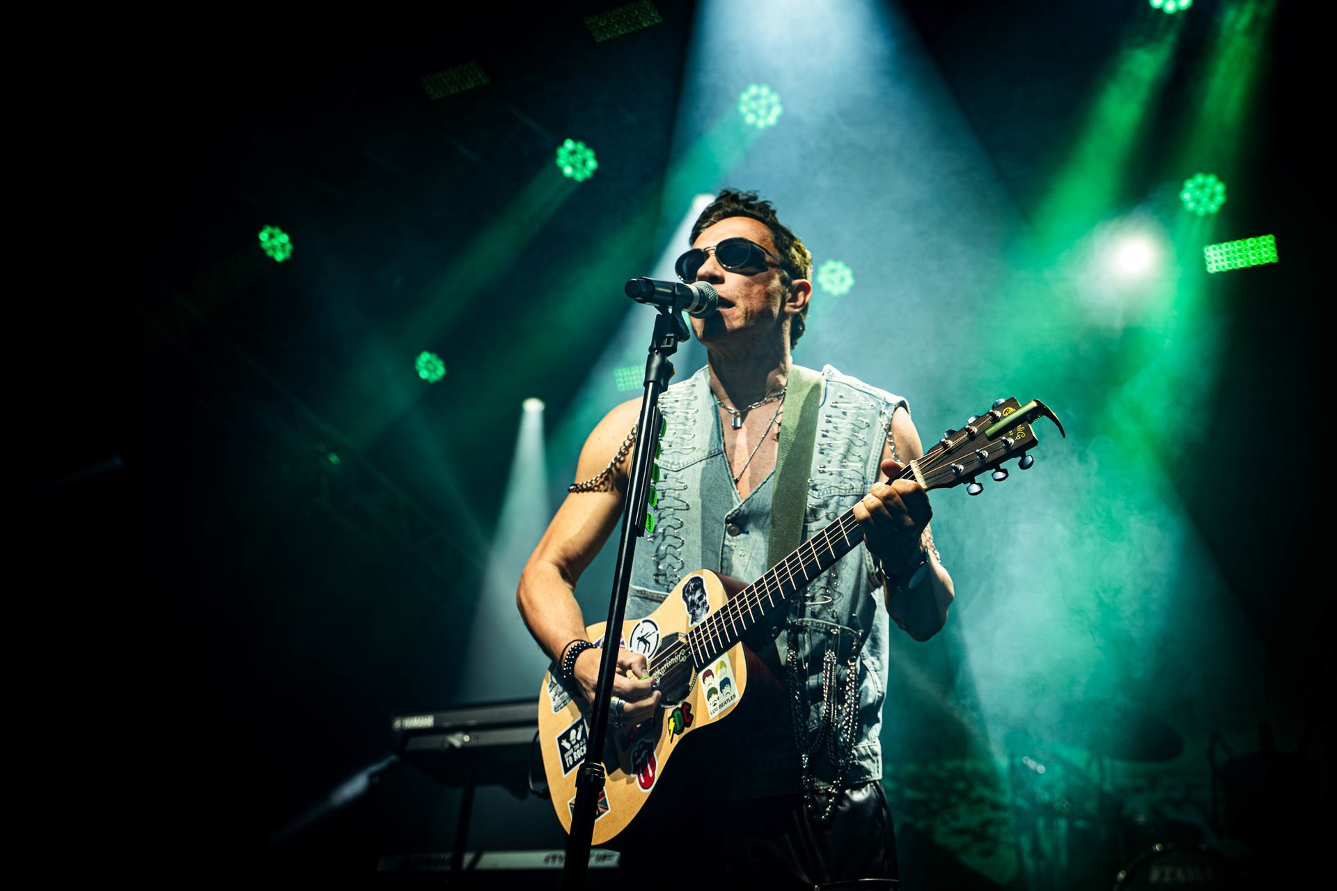 Guitarrista no palco, usando óculos escuros, tocando guitarra, cantando, palco iluminado com luzes verdes e brancas.