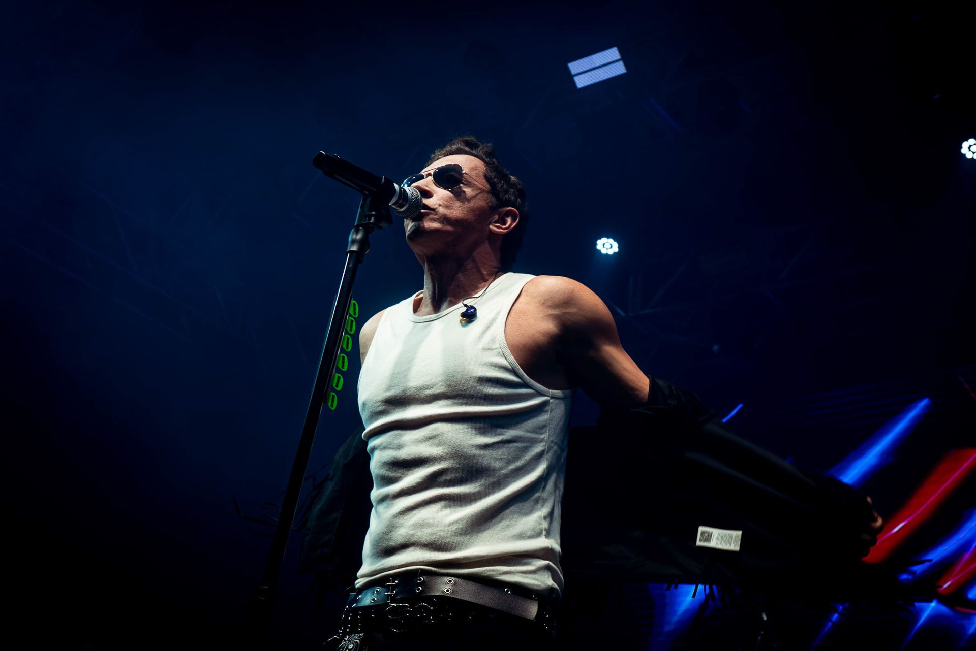 Um cantor de regata branca se apresenta no palco sob holofotes azuis, segurando um pedestal de microfone.