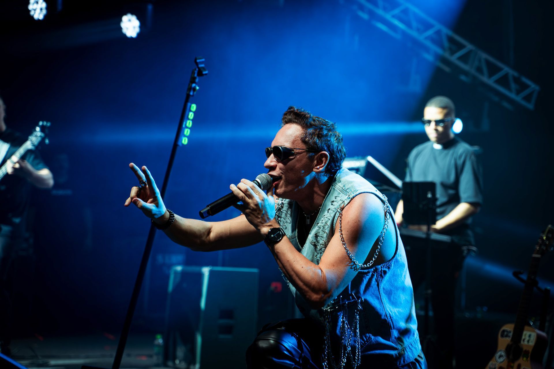 Um cantor de blusa sem mangas e óculos escuros se apresenta no palco sob luzes azuis, segurando um microfone e gesticulando.