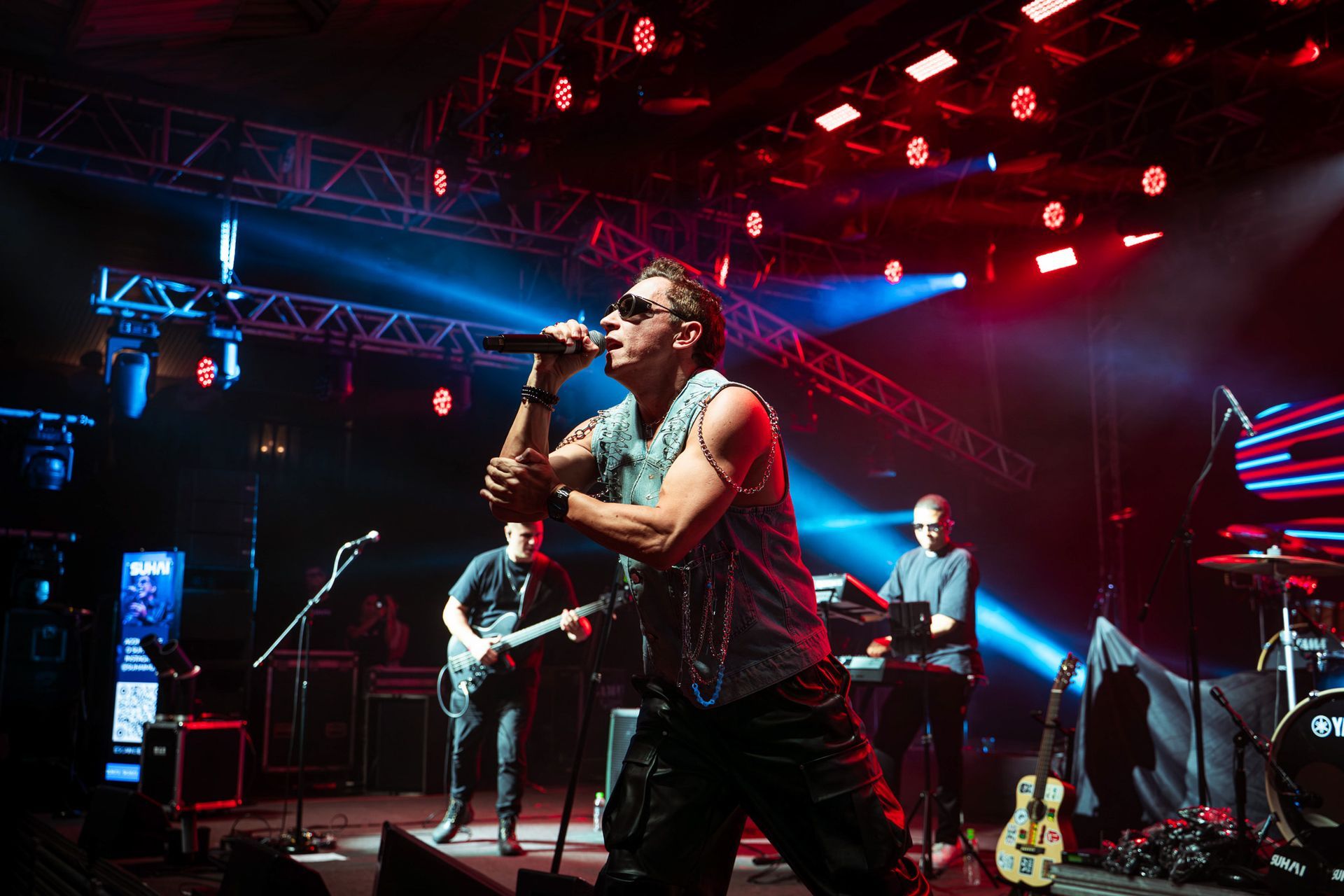 Um cantor de blusa sem mangas e óculos escuros se apresenta ao microfone em um palco com iluminação azul e vermelha de show.