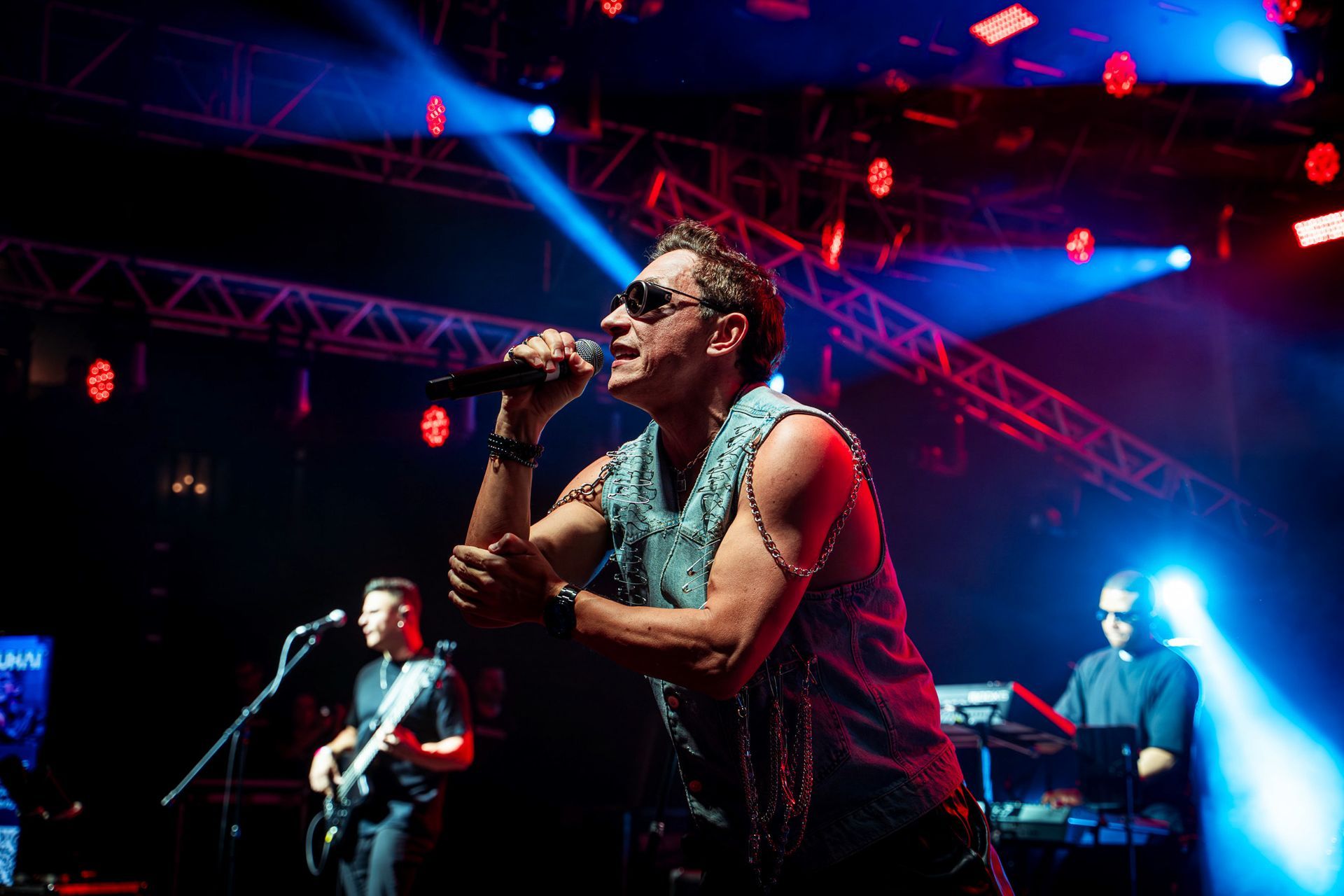Um cantor de colete jeans e óculos escuros se apresenta no palco sob luzes de concerto azuis e vermelhas