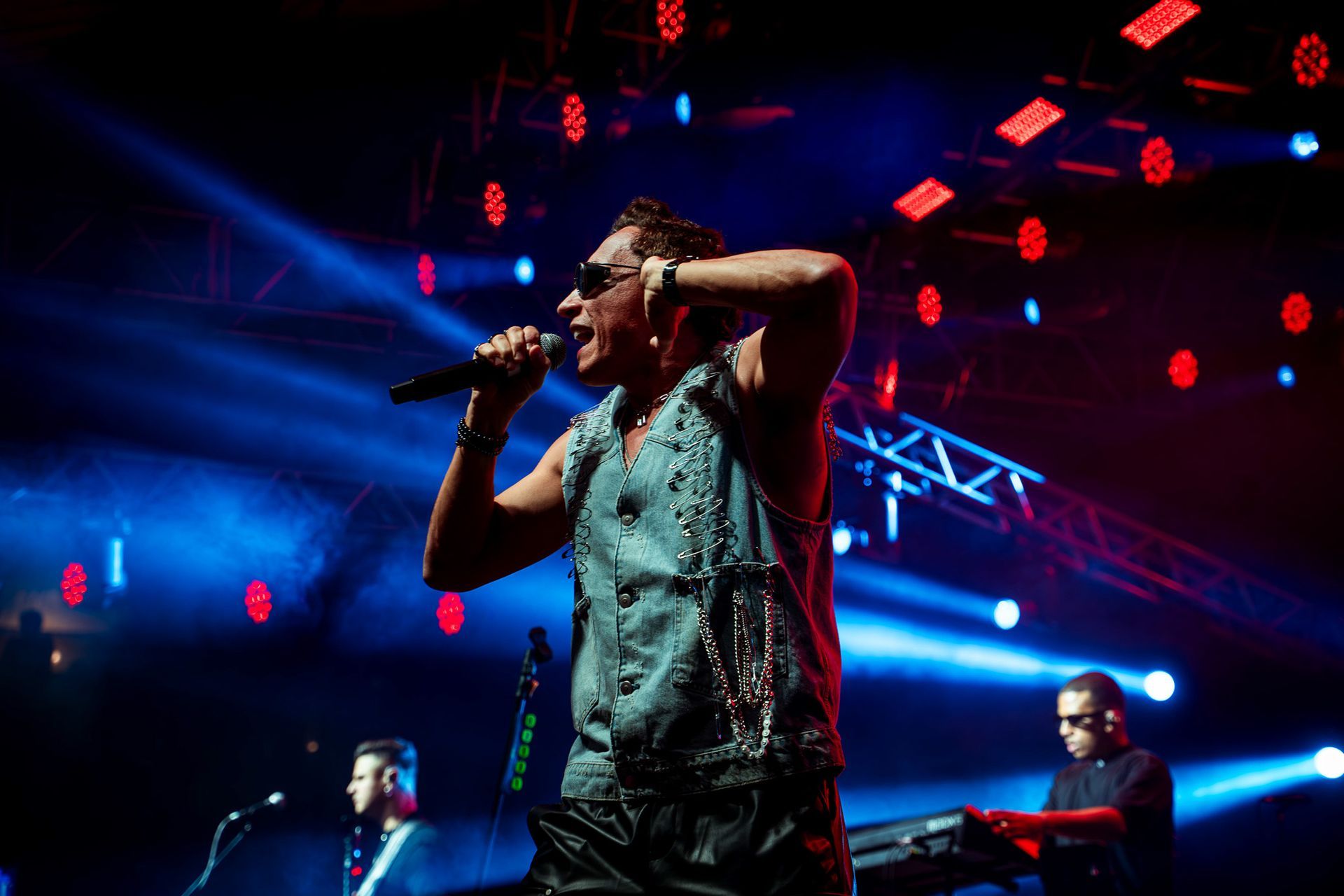 Um cantor vestindo colete e óculos escuros se apresenta em um palco iluminado por holofotes azuis, com um músico ao fundo.
