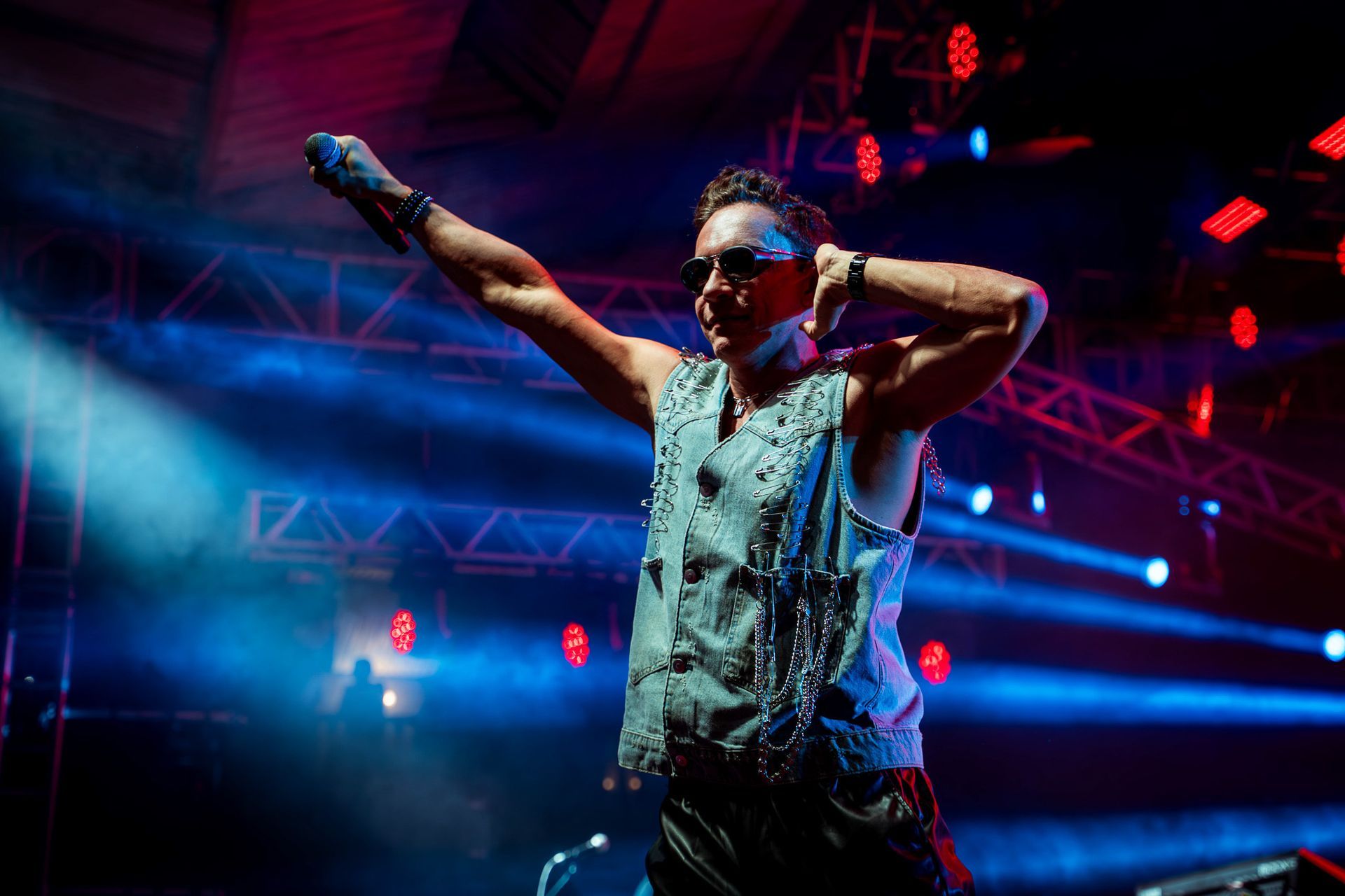 Um artista com um colete cravejado de tachas e óculos escuros estende a mão em um palco iluminado por holofotes azuis