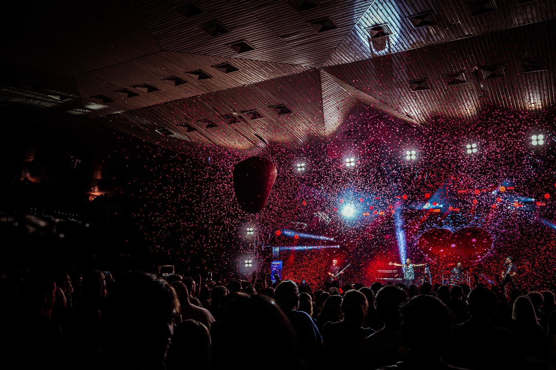 Um concerto com um palco iluminado em vermelho, artistas e uma multidão em silhueta e confetes vermelhos flutuantes.