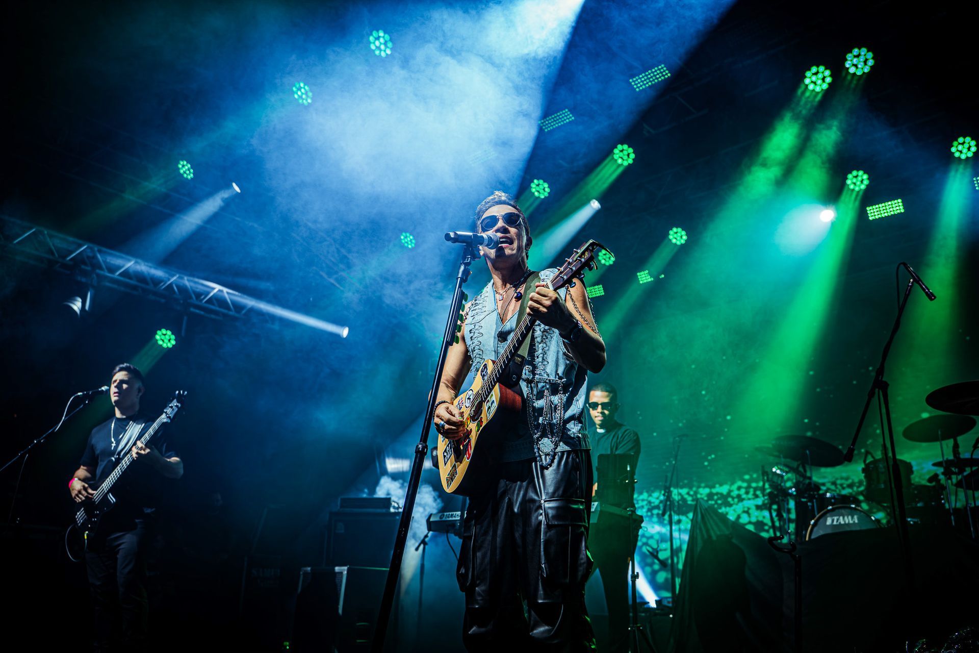 Uma banda se apresenta no palco sob luzes azuis e verdes. O vocalista toca guitarra e canta.