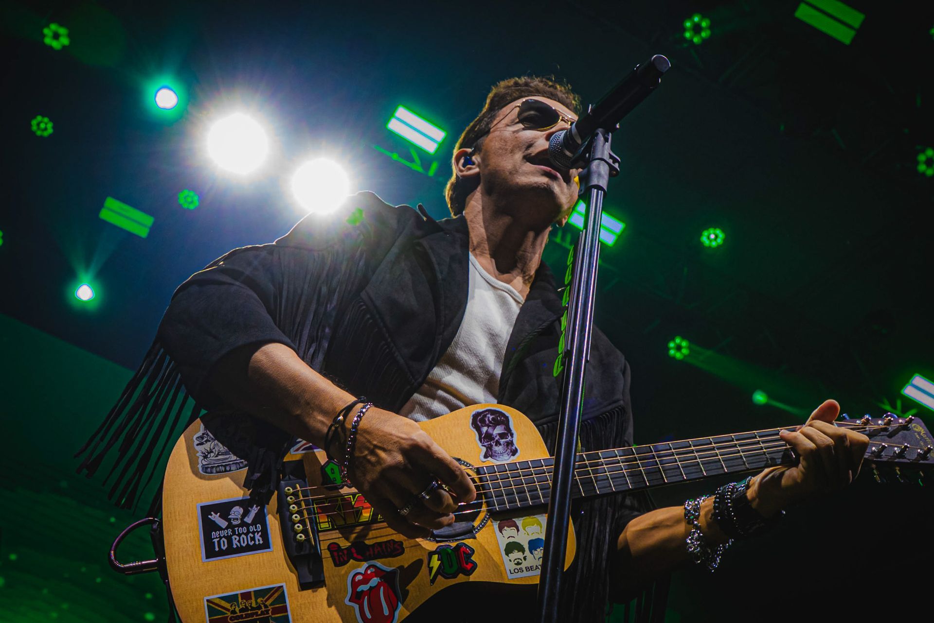 Um músico com um violão coberto de adesivos se apresenta em um palco iluminado por holofotes verdes brilhantes.