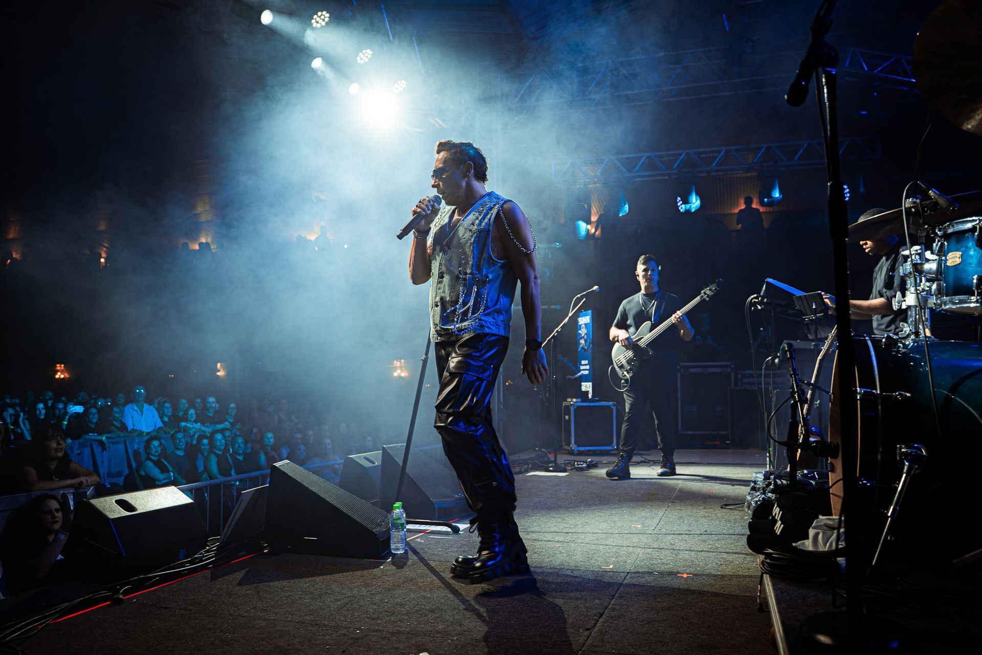 Um vocalista se apresenta no palco com um guitarrista e um baterista, sob a iluminação dramática do palco