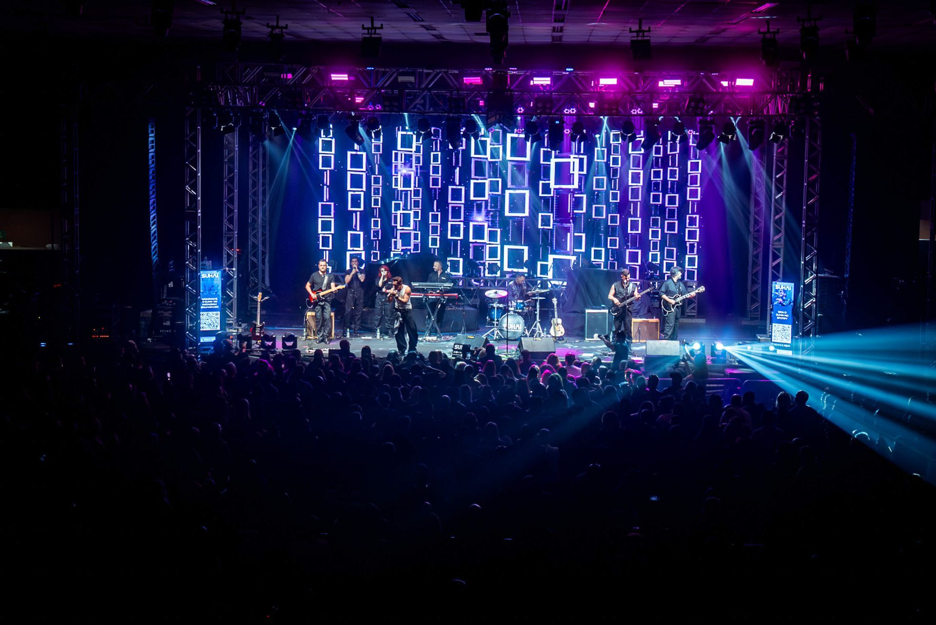Uma banda se apresenta em um palco banhado por luzes azuis e roxas flutuantes e iluminadas.