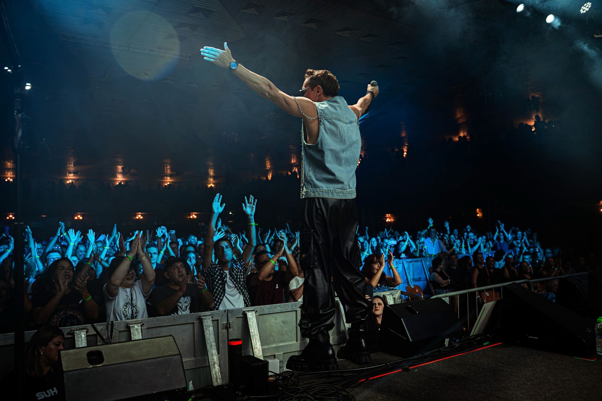 Um artista vestindo um colete jeans está em um palco com os braços estendidos diante de uma plateia entusiasmada 