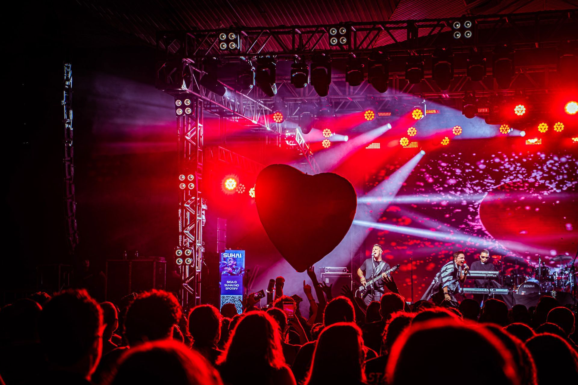 Uma banda se apresenta em um palco banhado por luzes vermelhas, balão em forma de coração flutuando acima da plateia.