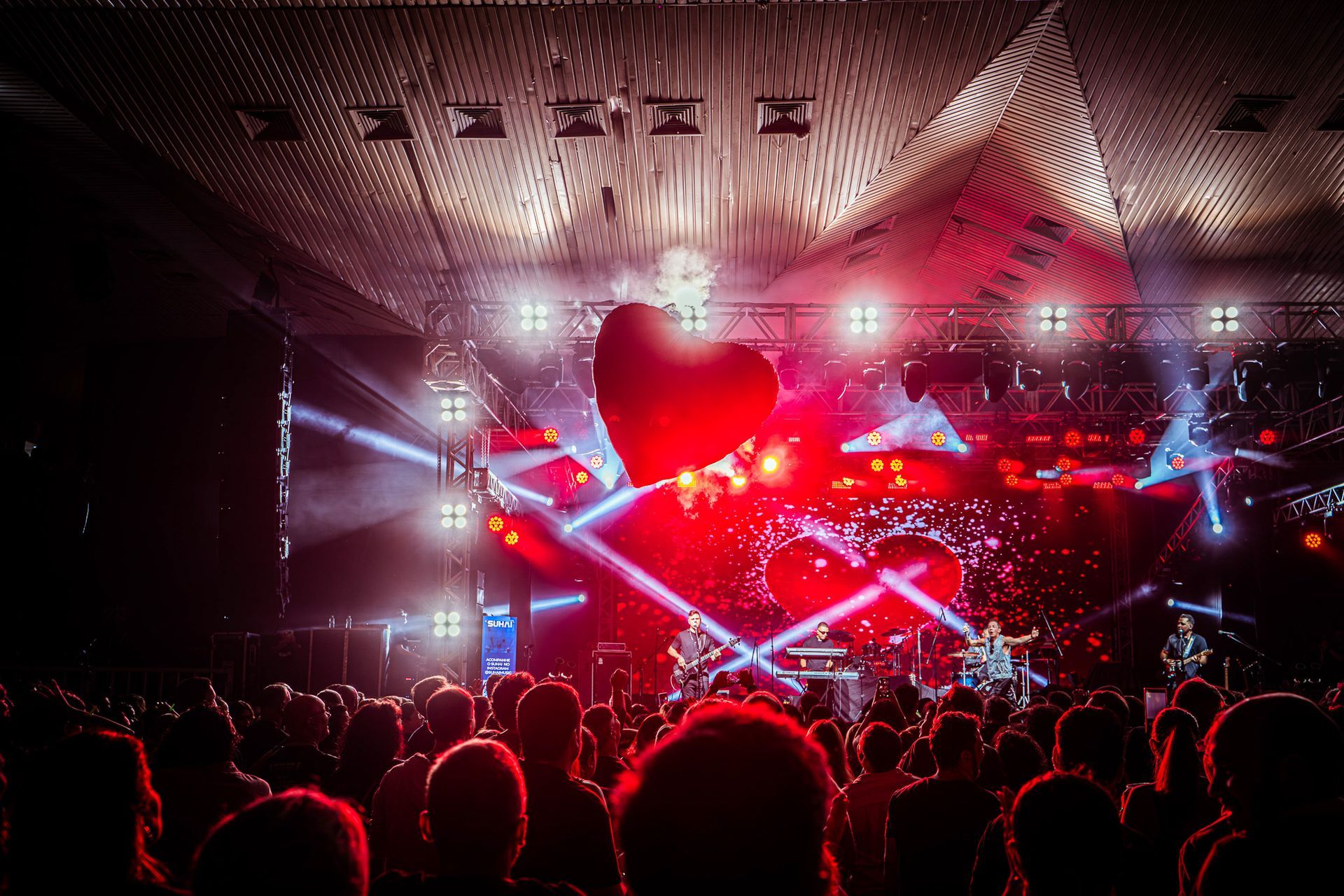 Uma apresentação de concerto ao vivo com uma grande decoração em forma de coração vermelho flutua em frente a uma plateia.