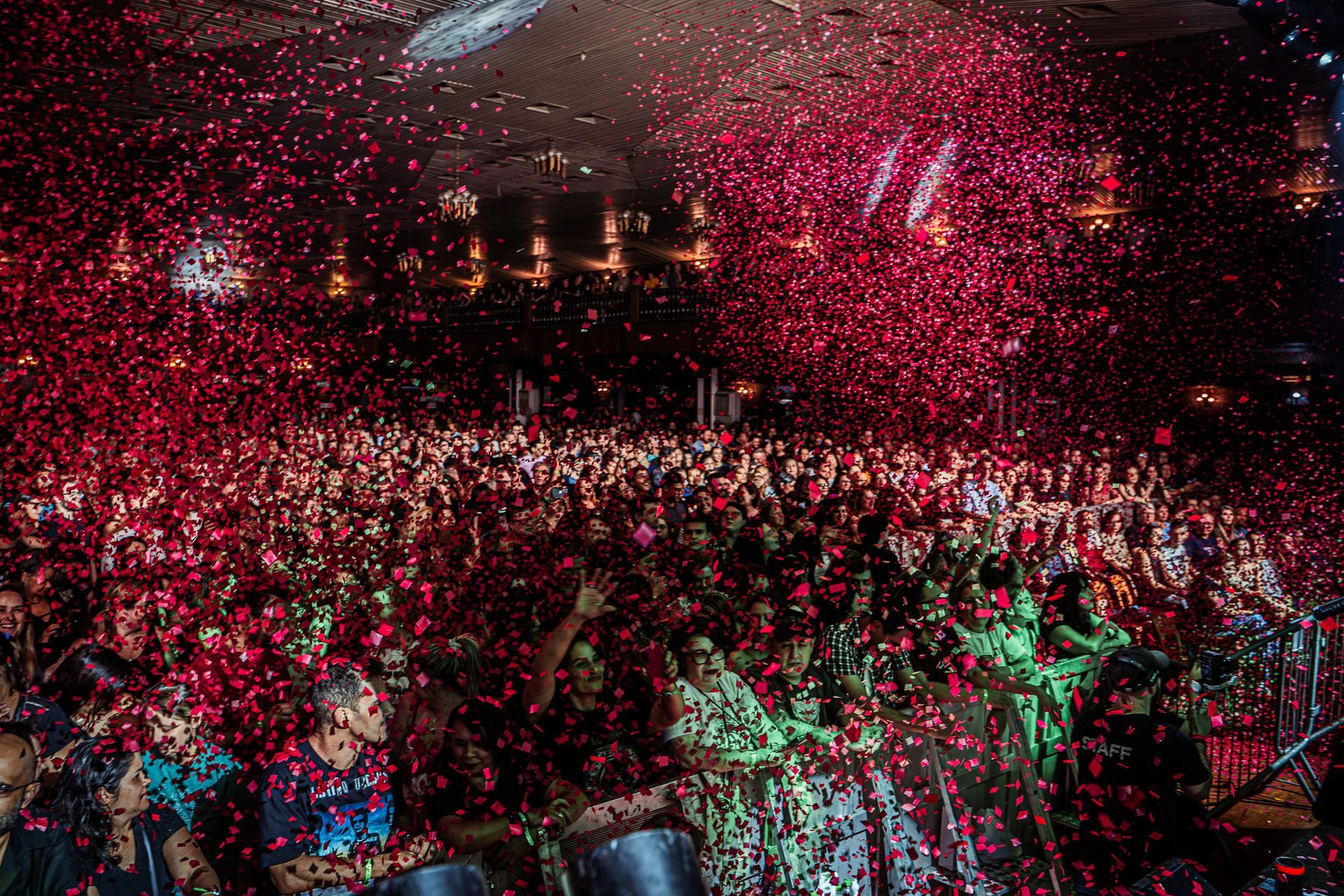O público assiste a um concerto em recinto fechado enquanto confetes vermelhos caem por toda a sala.