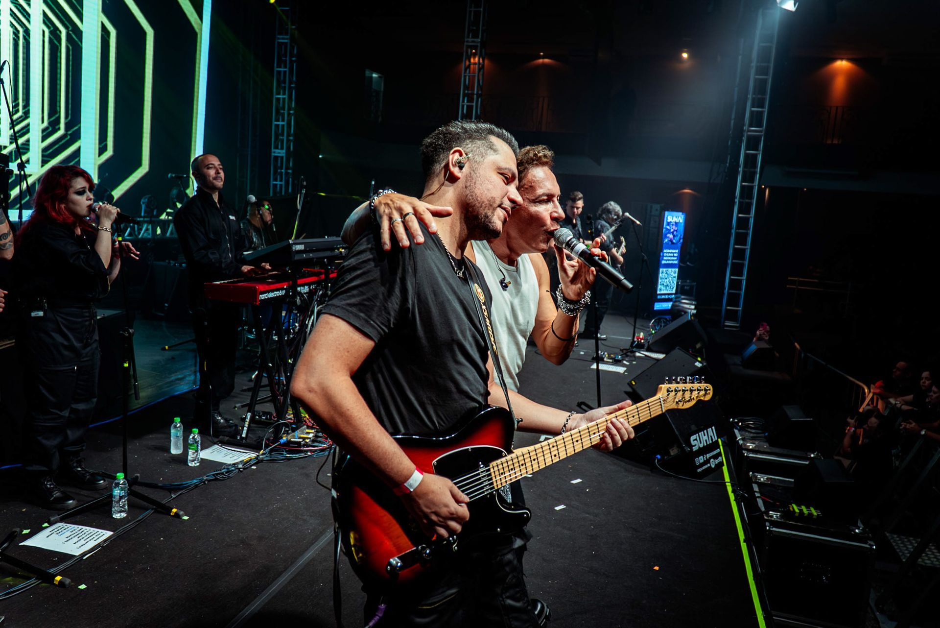 Dois artistas em um palco de concerto com pouca luz, um cantando ao microfone e o outro tocando guitarra elétrica.
