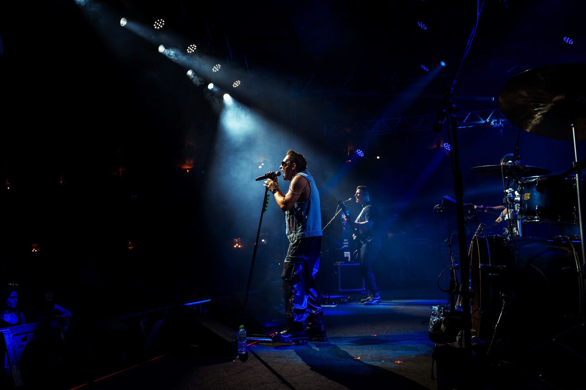 Um cantor se apresenta em um palco escuro, banhada por um dramático holofote azul, com um baterista visível ao fundo.