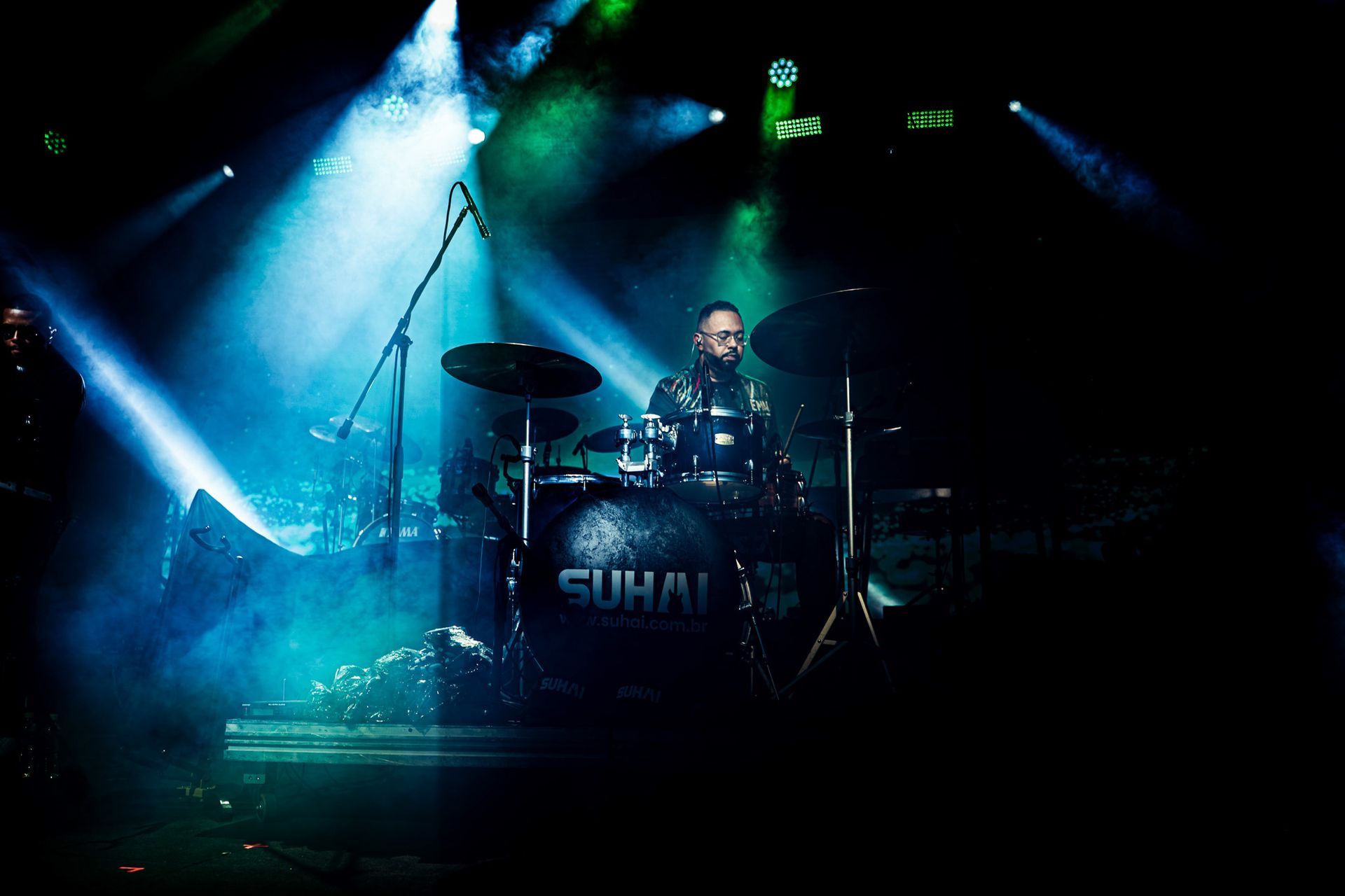 Baterista no palco, iluminado por holofotes azuis e verdes, tocando atrás de uma bateria com o nome 