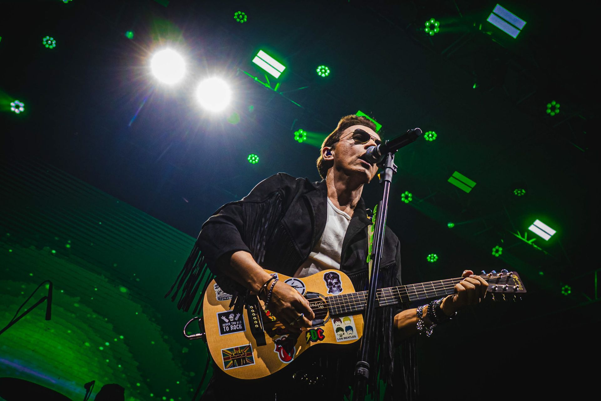 Um artista tocando um violão coberto de adesivos e cantando em um microfone sob luzes de palco verde-brilhantes.