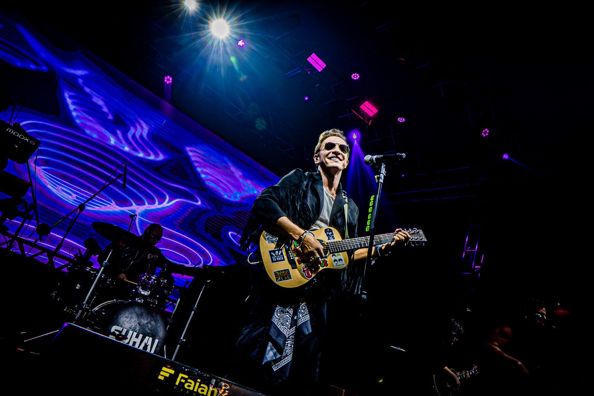 Um artista de óculos escuros sorri enquanto toca violão em um palco escuro com efeitos de luz azul brilhante.