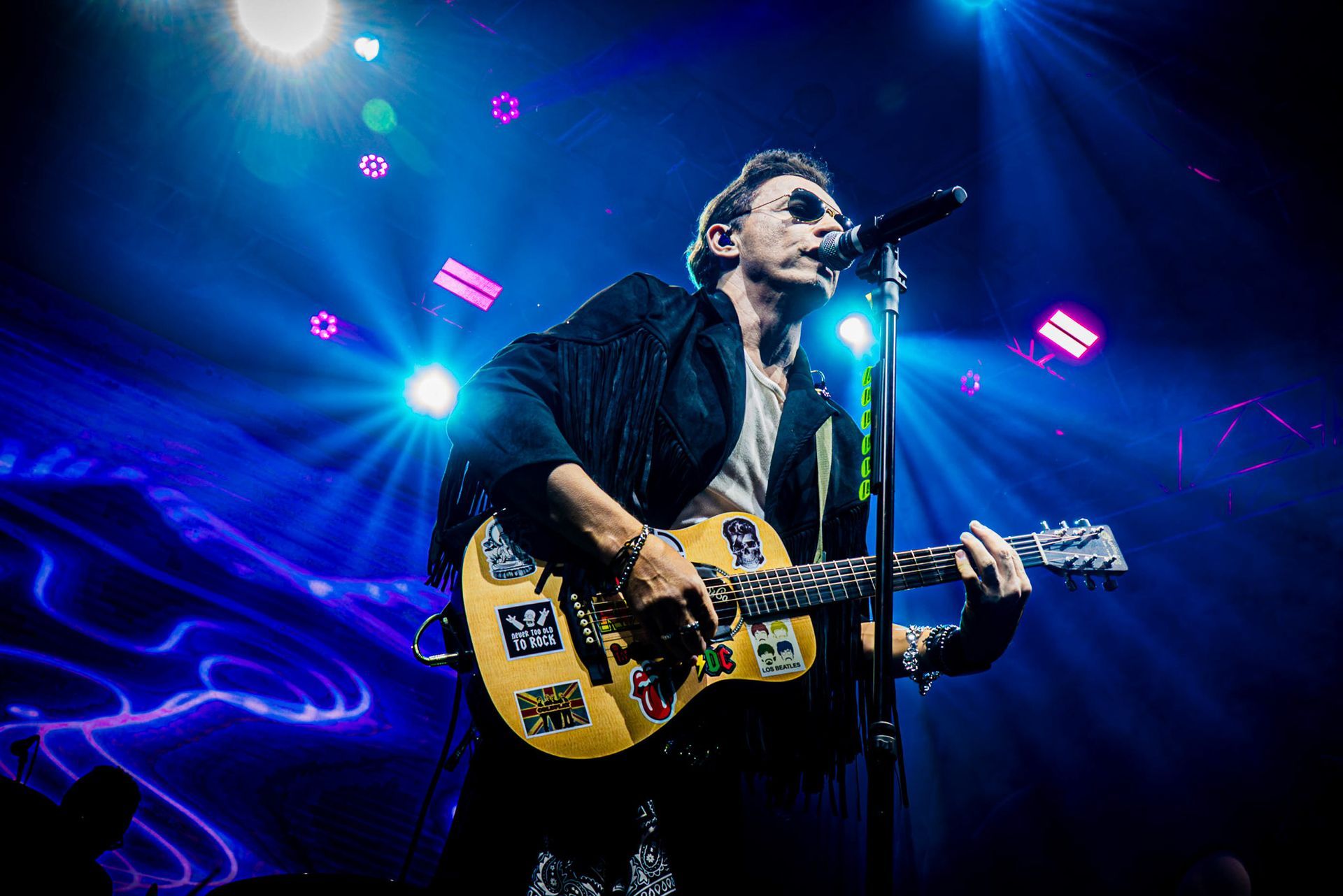 Um músico usando óculos escuros toca um violão coberto de adesivos enquanto canta em um microfone no palco.