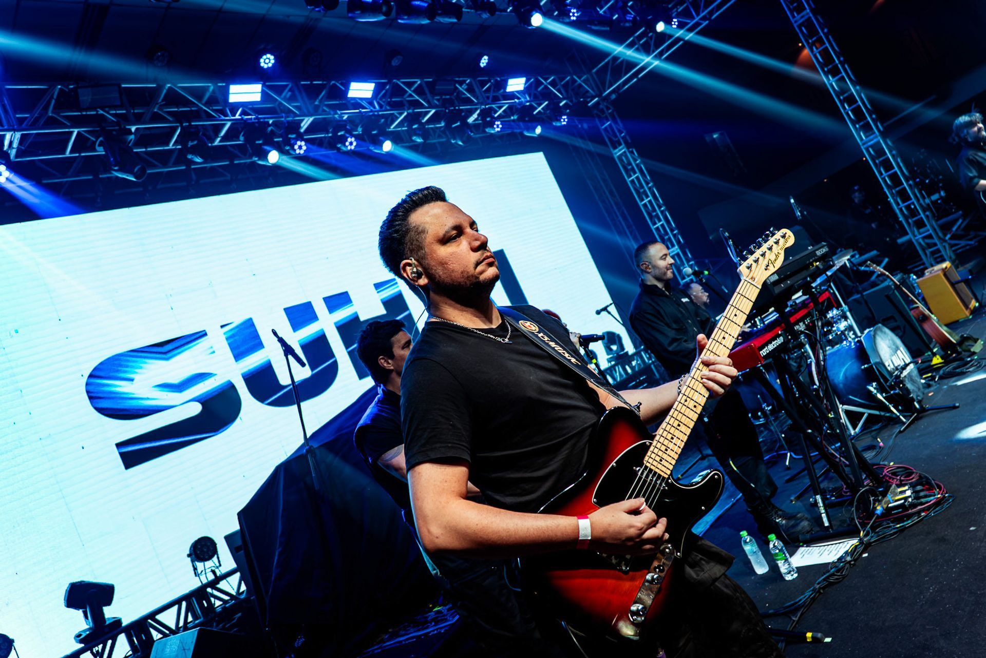 Um músico toca uma guitarra elétrica vermelha em um palco de concertos com luzes de palco brilhantes e um telão ao fundo.