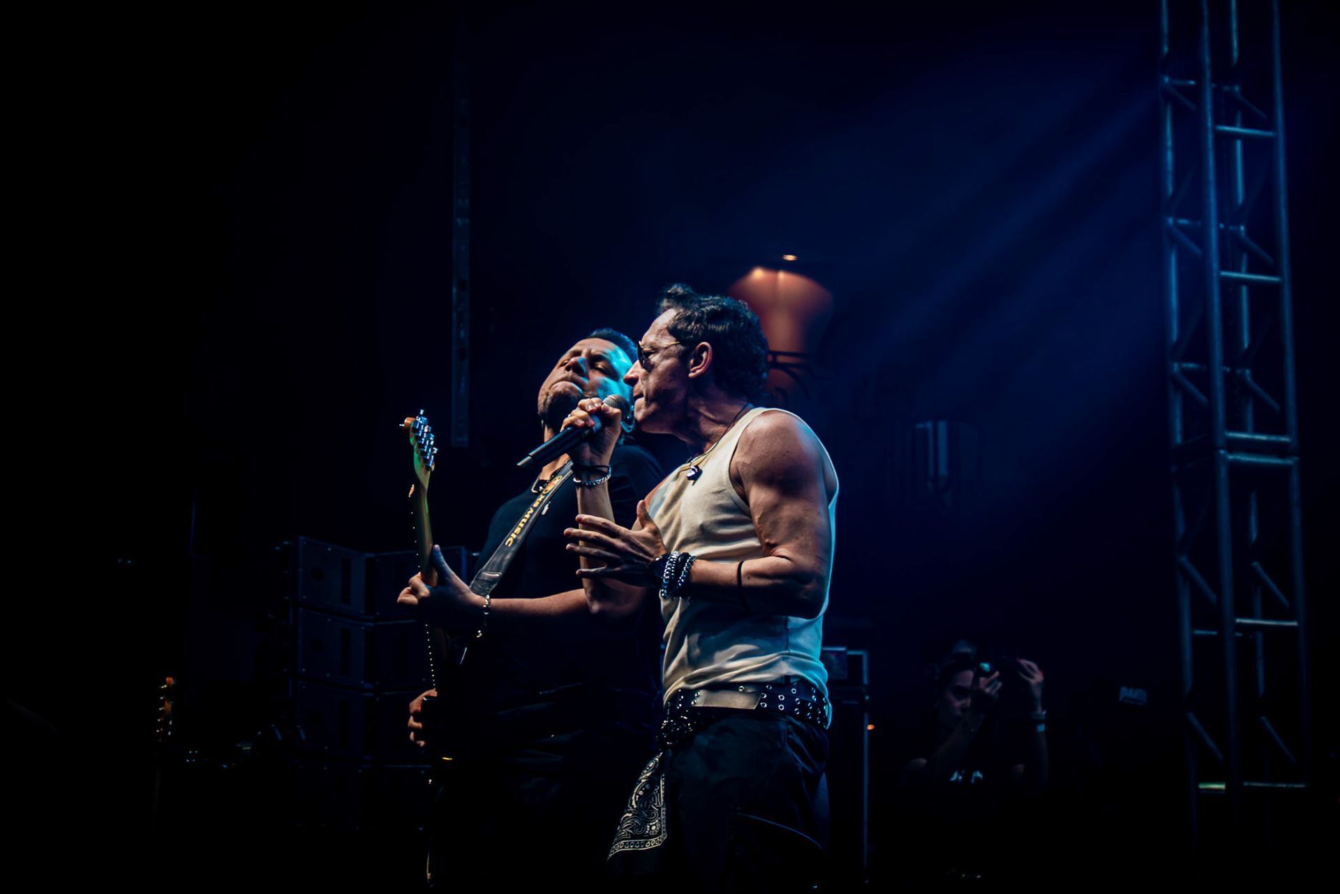 Dois artistas em um palco com pouca luz, um cantando ao microfone enquanto o outro toca guitarra elétrica.