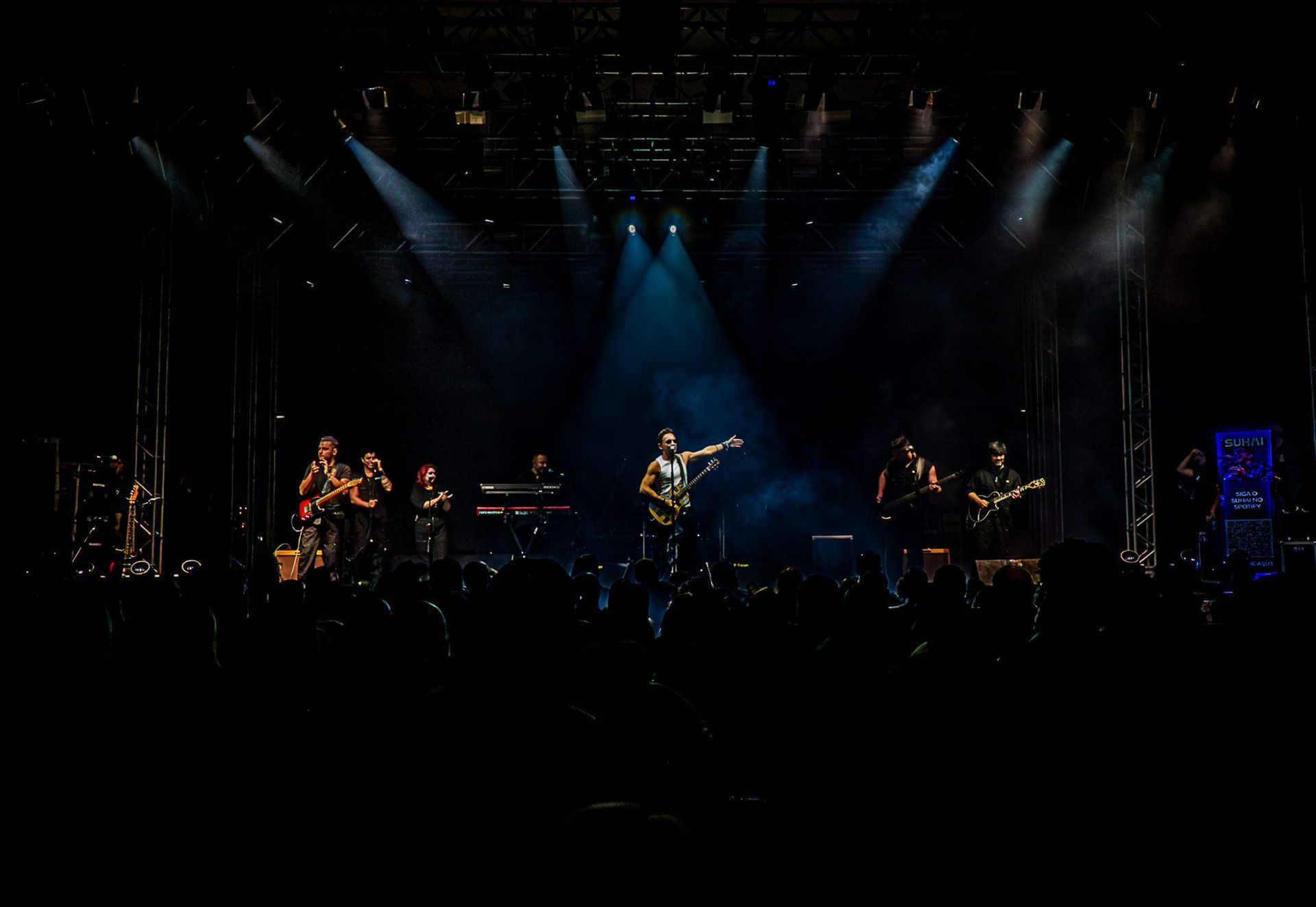 Uma banda se apresenta em um palco escuro, iluminado por fortes holofotes azuis, com o vocalista gesticulando para a plateia.