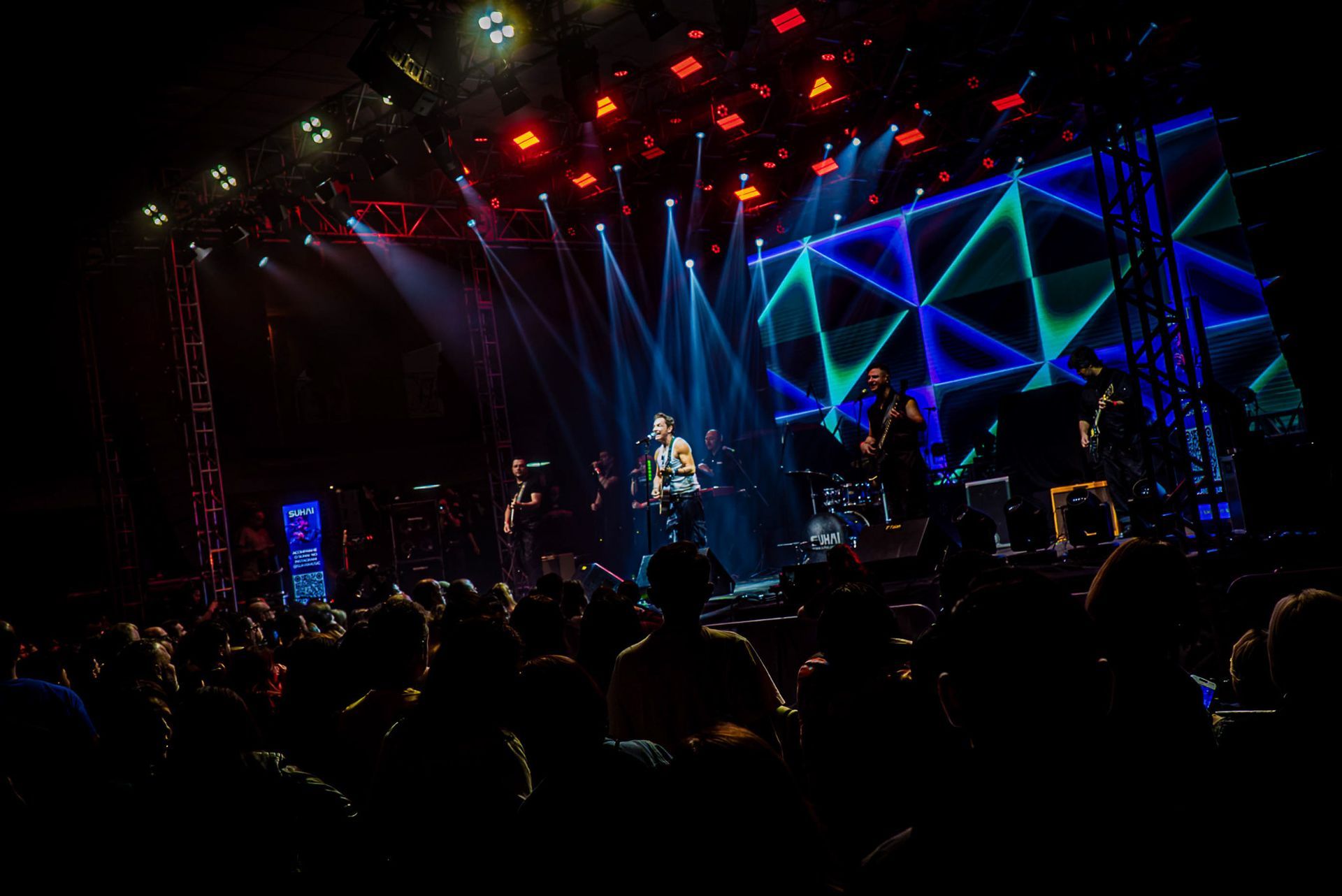 Um concerto ao vivo à noite com um músico a atuar num palco iluminado por holofotes azuis brilhantes e um cenário geométrico.